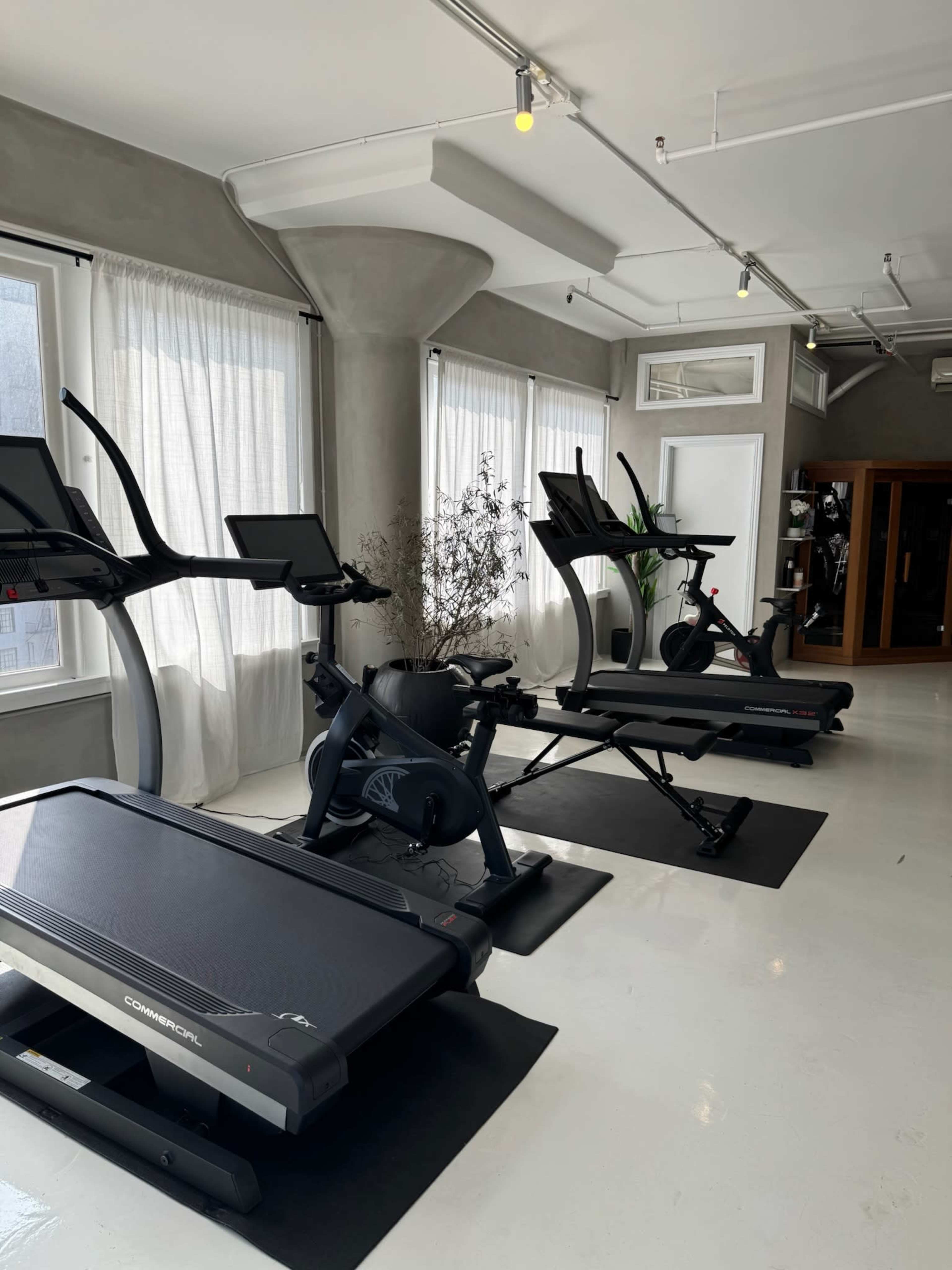 The image shows a modern gym interior with various exercise equipment including treadmills and elliptical trainers, placed in front of large windows with natural light.