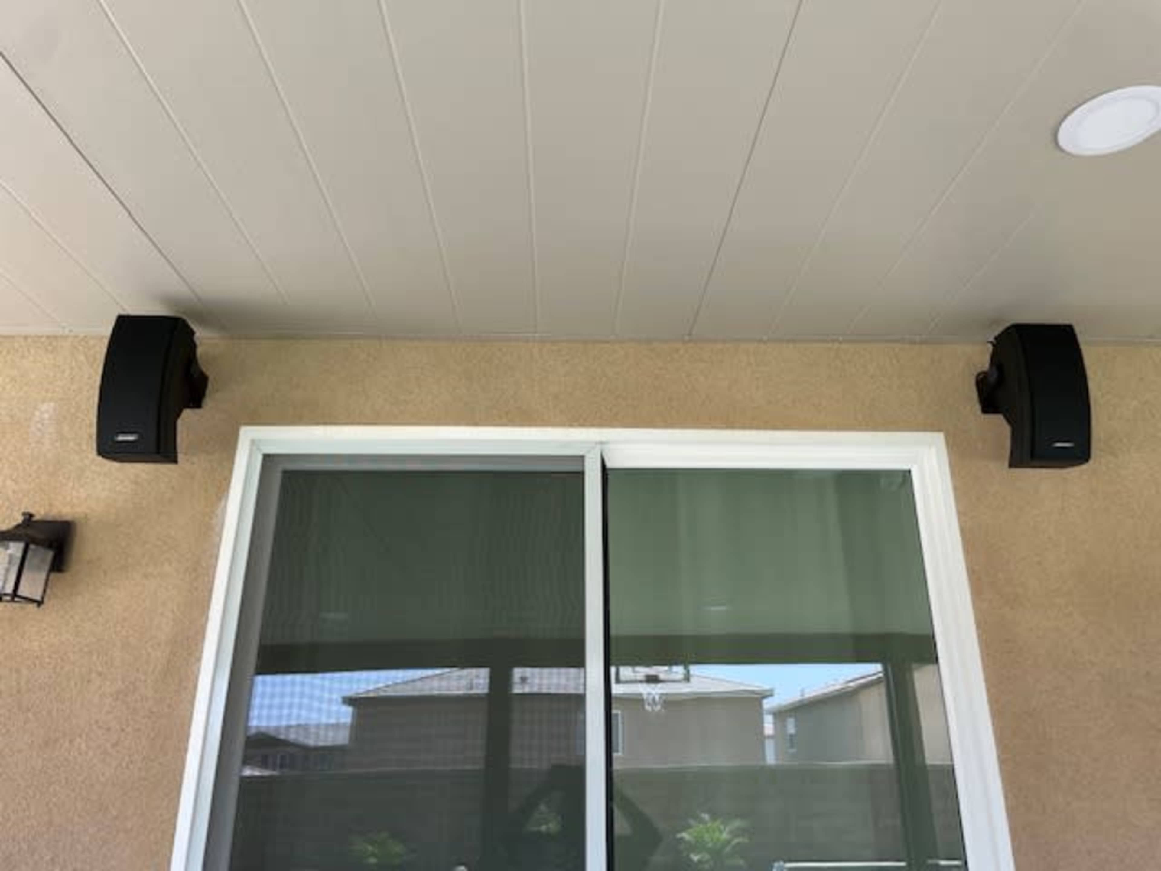 The image shows two black speakers mounted on the wall above a large sliding glass door.