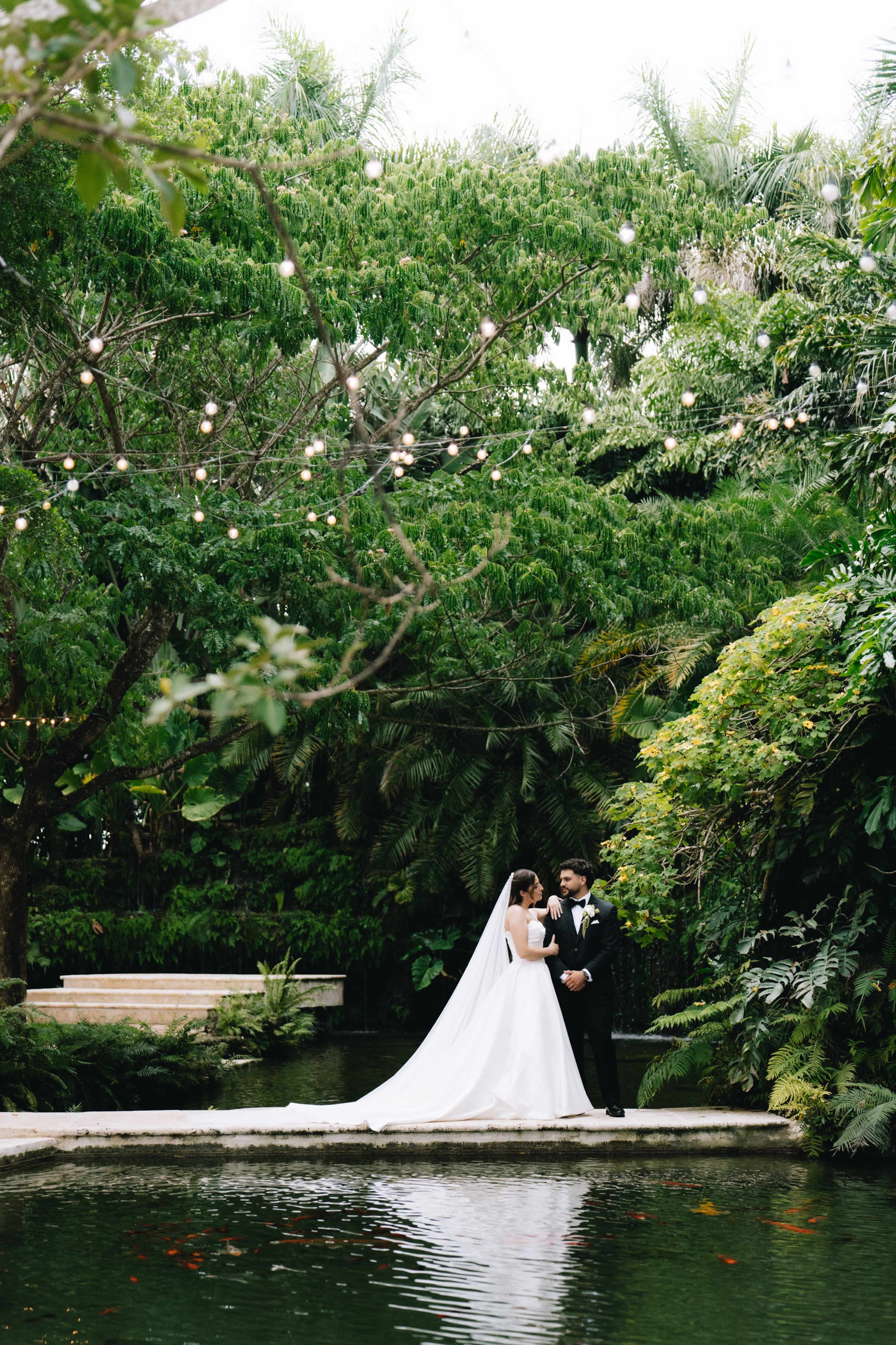 A couple in formal wedding attire stands together on a path over a tranquil pond surrounded by lush greenery and string lights.