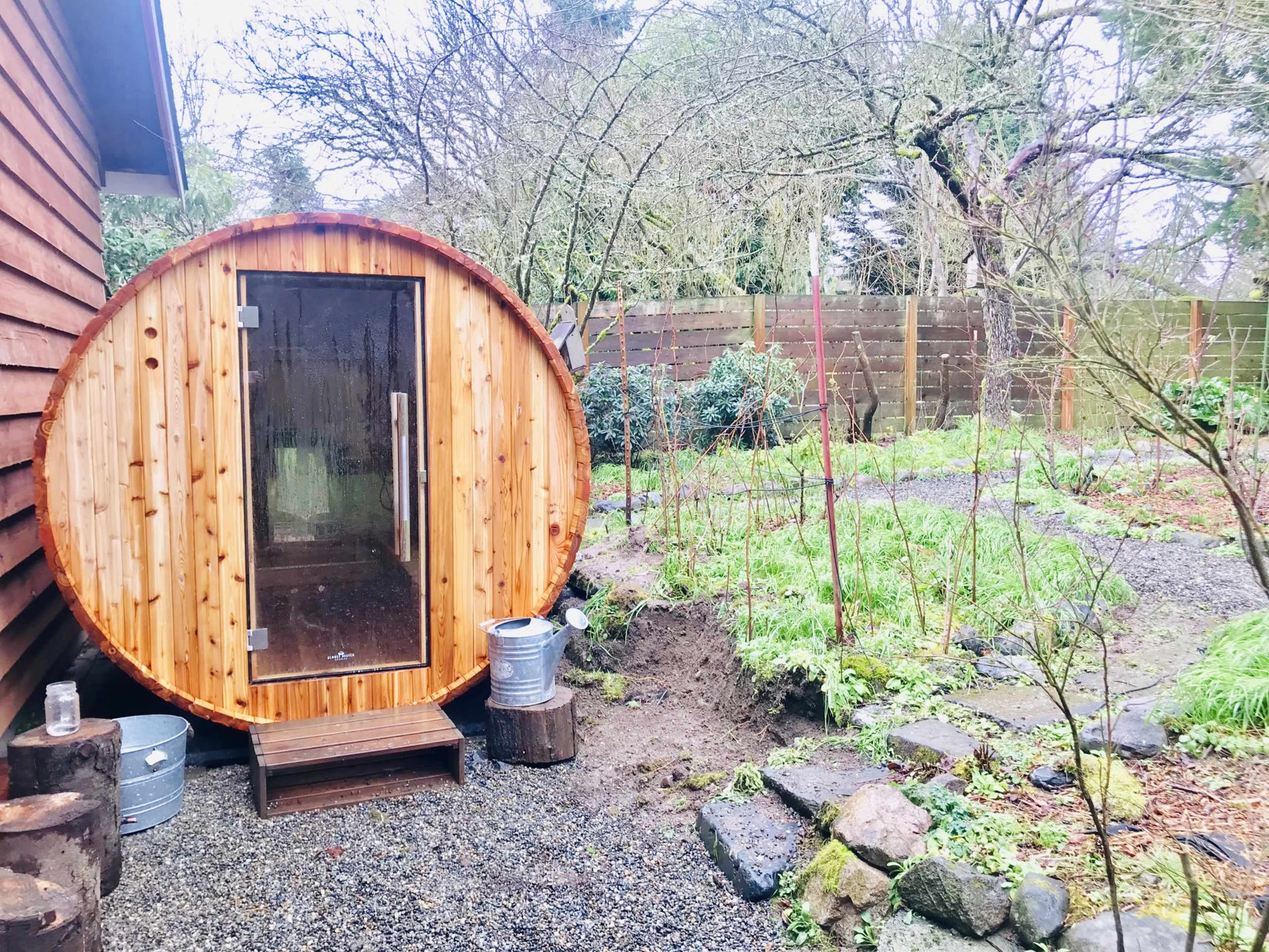 A round wooden sauna situated next to a gravel path in a garden with greenery and trees in the background.