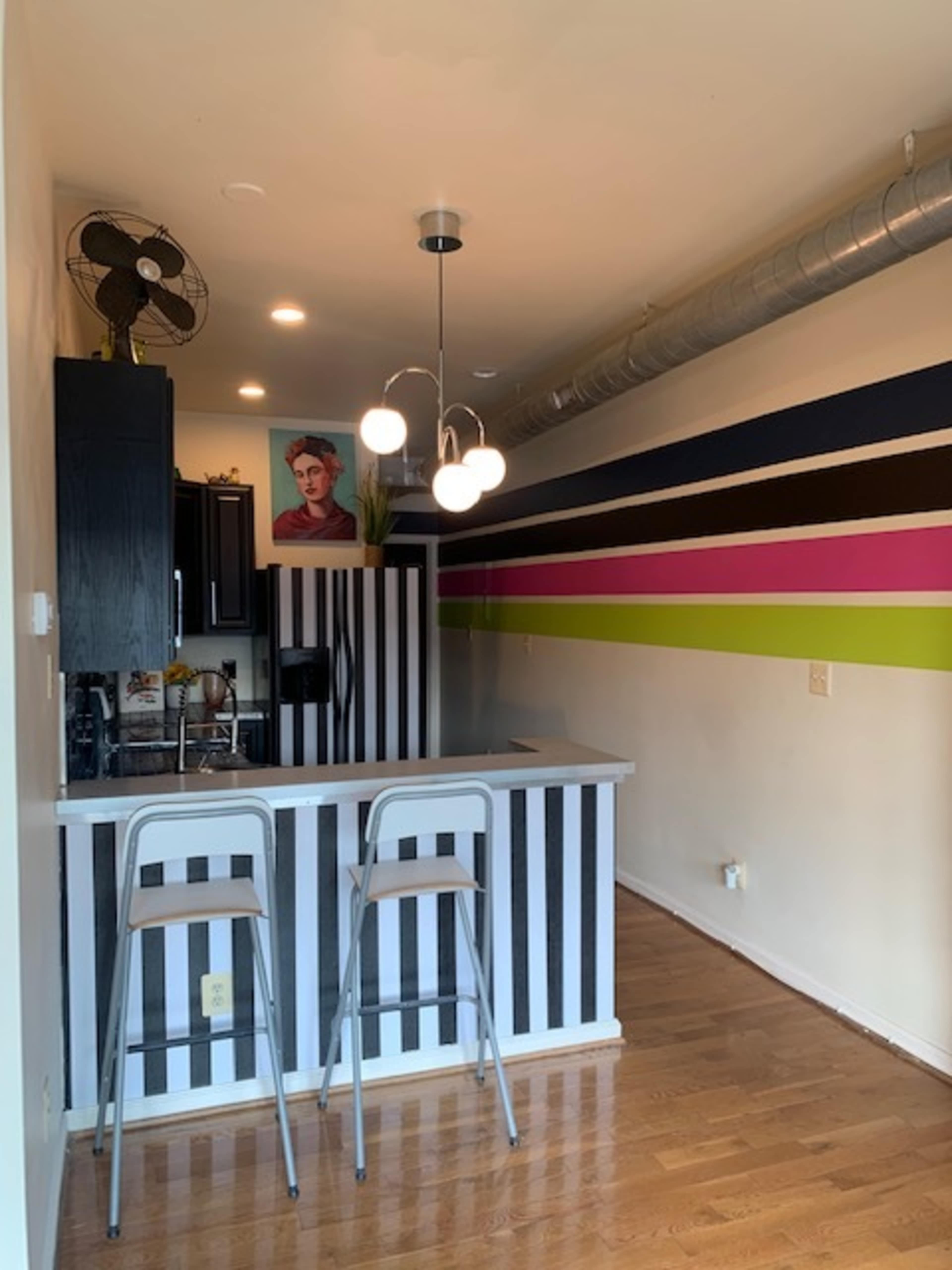 The image shows a modern kitchen with a bar area featuring striped black and white panels, two metal stools, and colorful horizontal wall stripes.