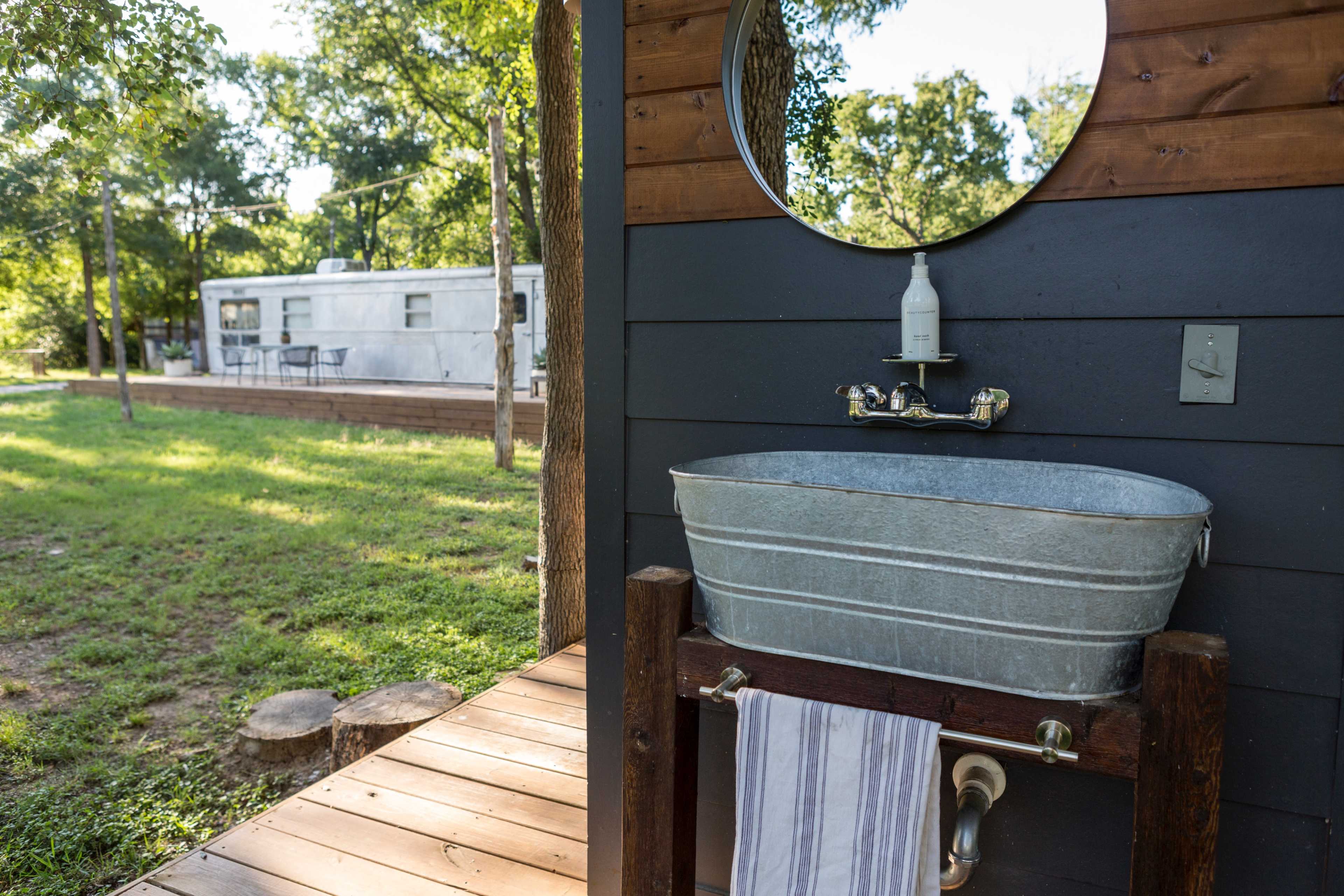 An outdoor sink made from a galvanized tub is positioned on a wooden deck beside a grassy area, with a silver trailer visible in the background.