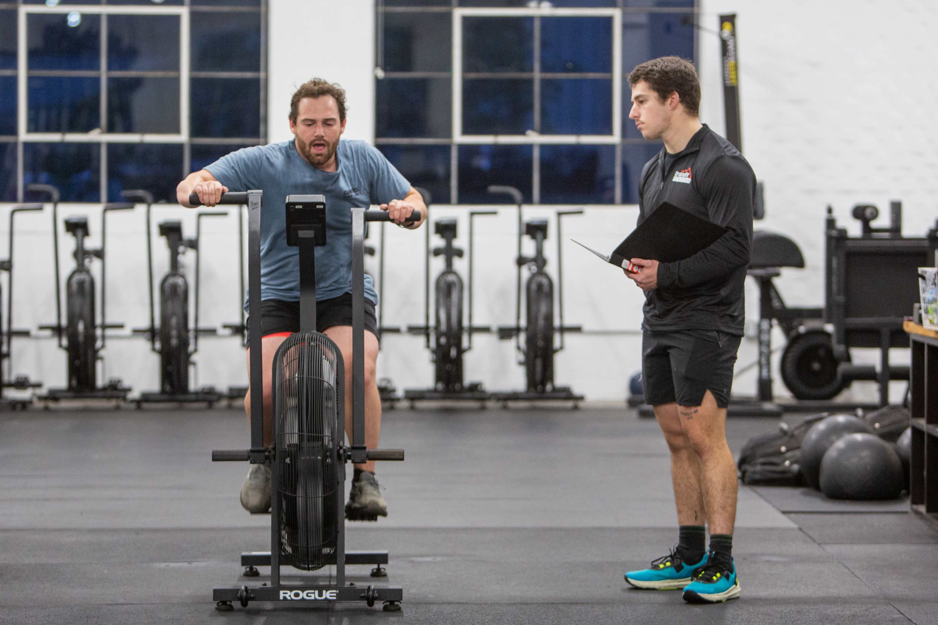 A man exercises on a rowing machine while another man observes and holds a clipboard nearby in a gym setting.