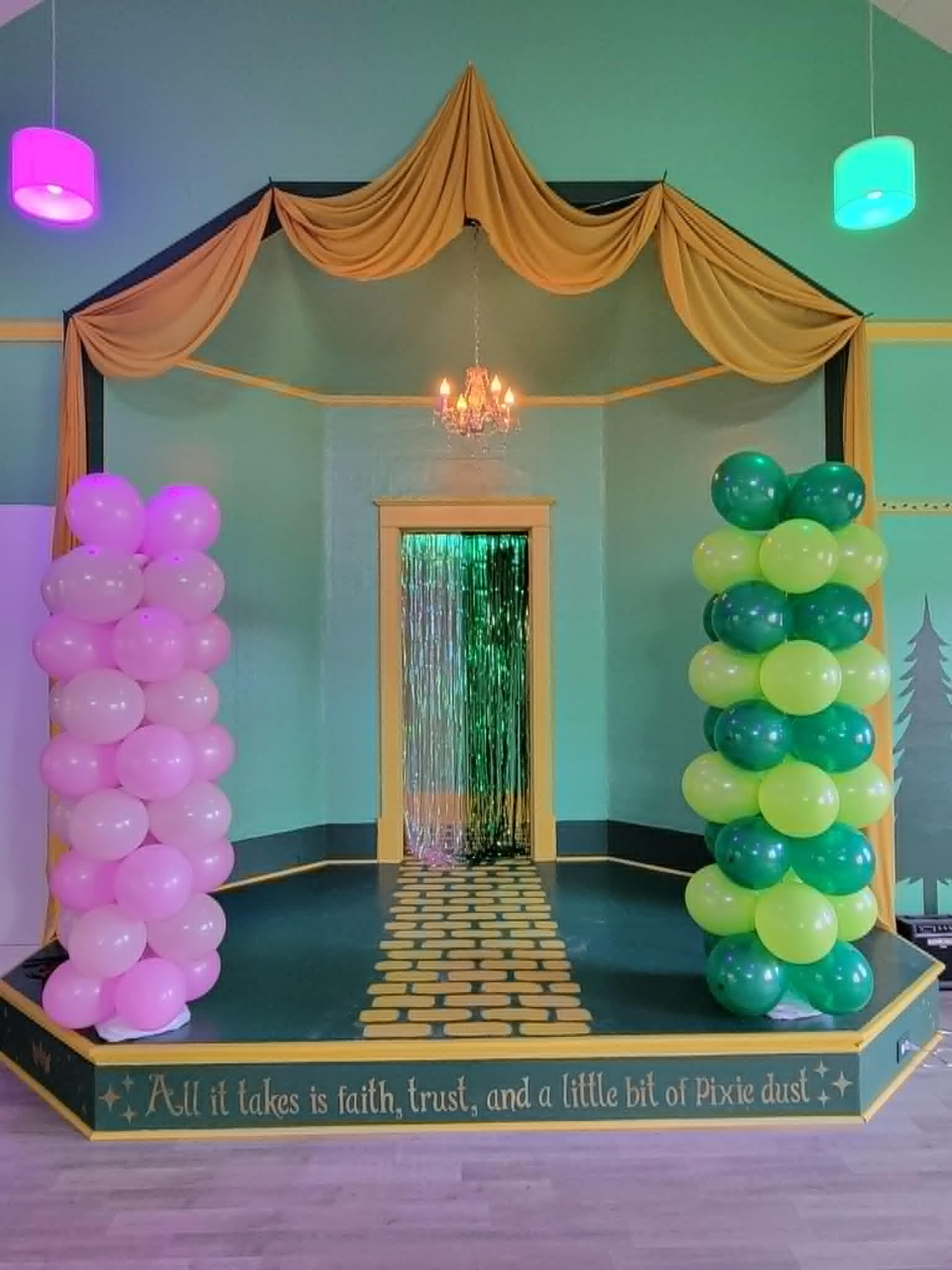 The image shows a decorated stage with pink and green balloon columns, a shimmering curtain at the back, and a quote displayed along the bottom.