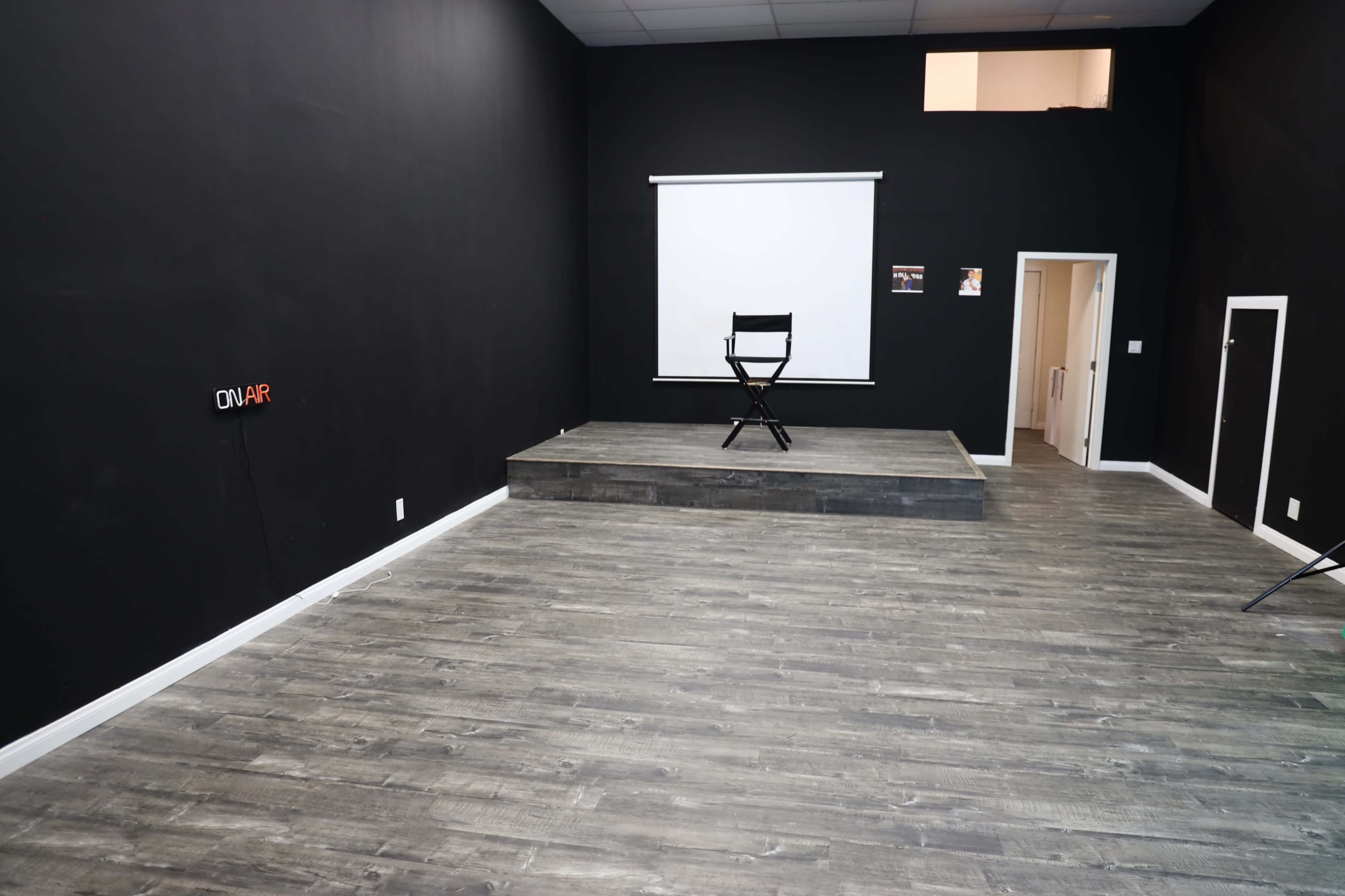 The image shows a large, empty black room with a raised platform featuring a single black chair, a projection screen, and a doorway in the background.