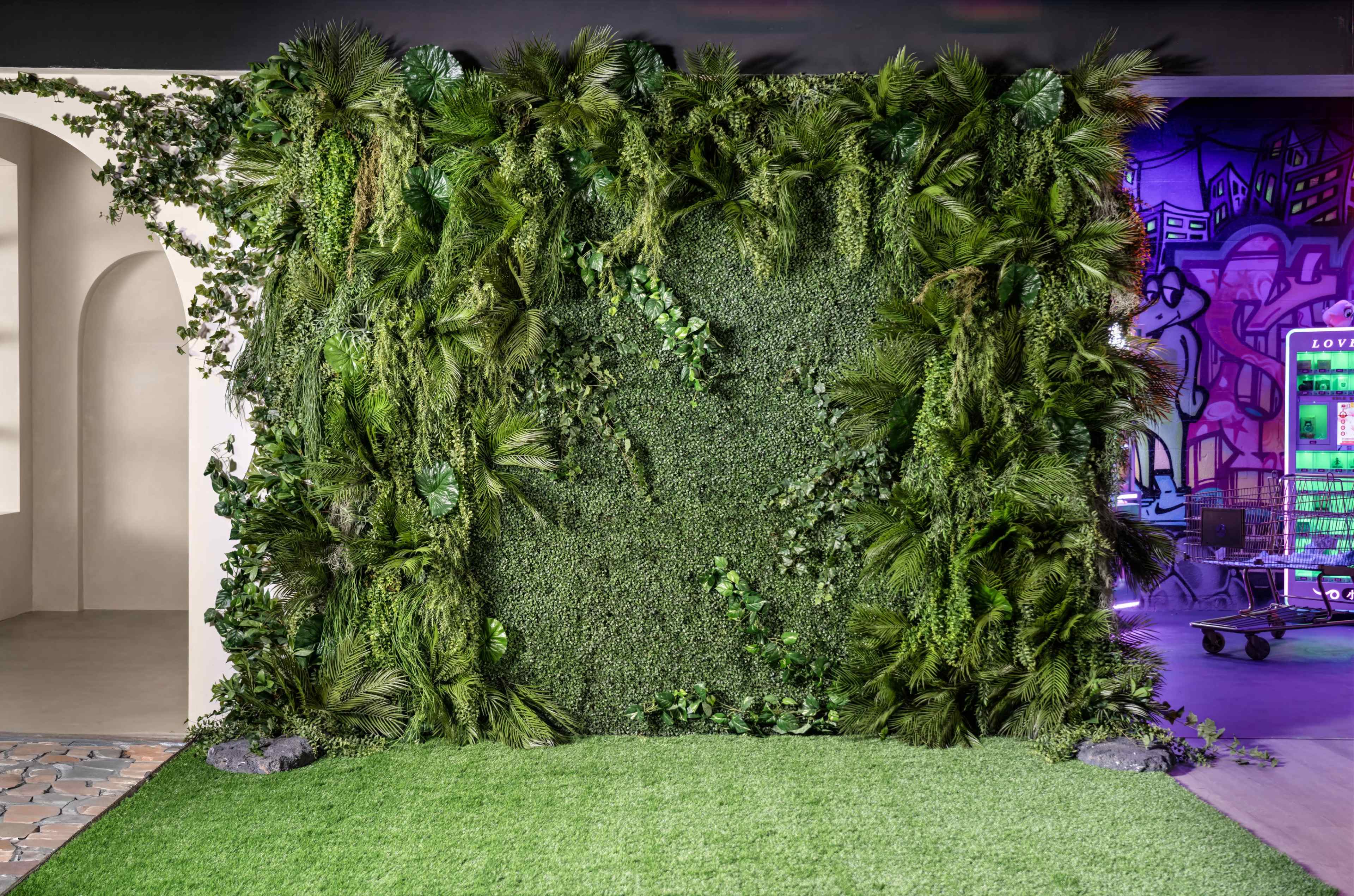 A large green wall of artificial plants and foliage, set against a backdrop with a hint of illuminated urban artwork.