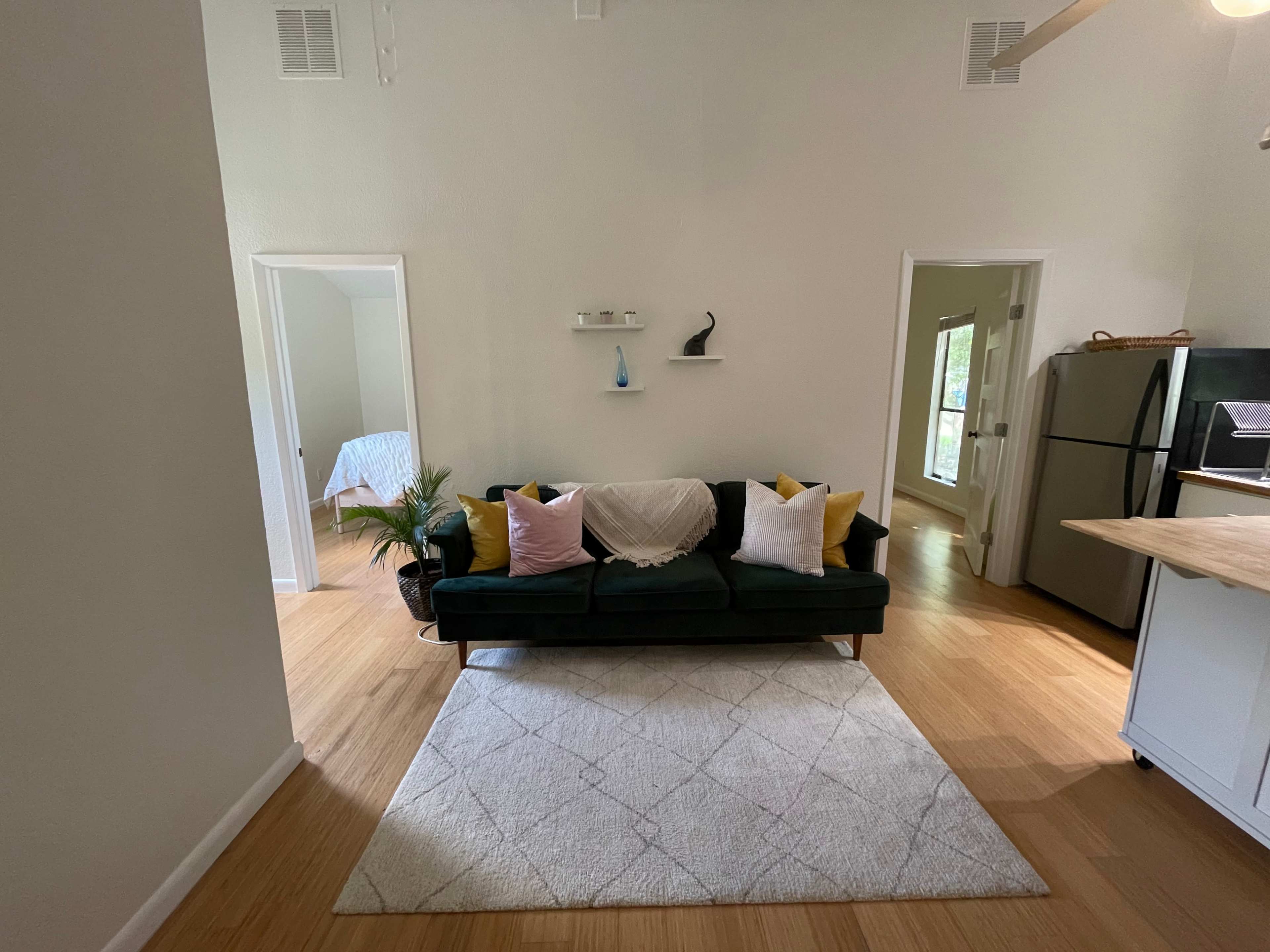 A living room with a green sofa adorned with colorful pillows, a light rug on a wooden floor, and an open layout leading to a kitchen and a bedroom area.