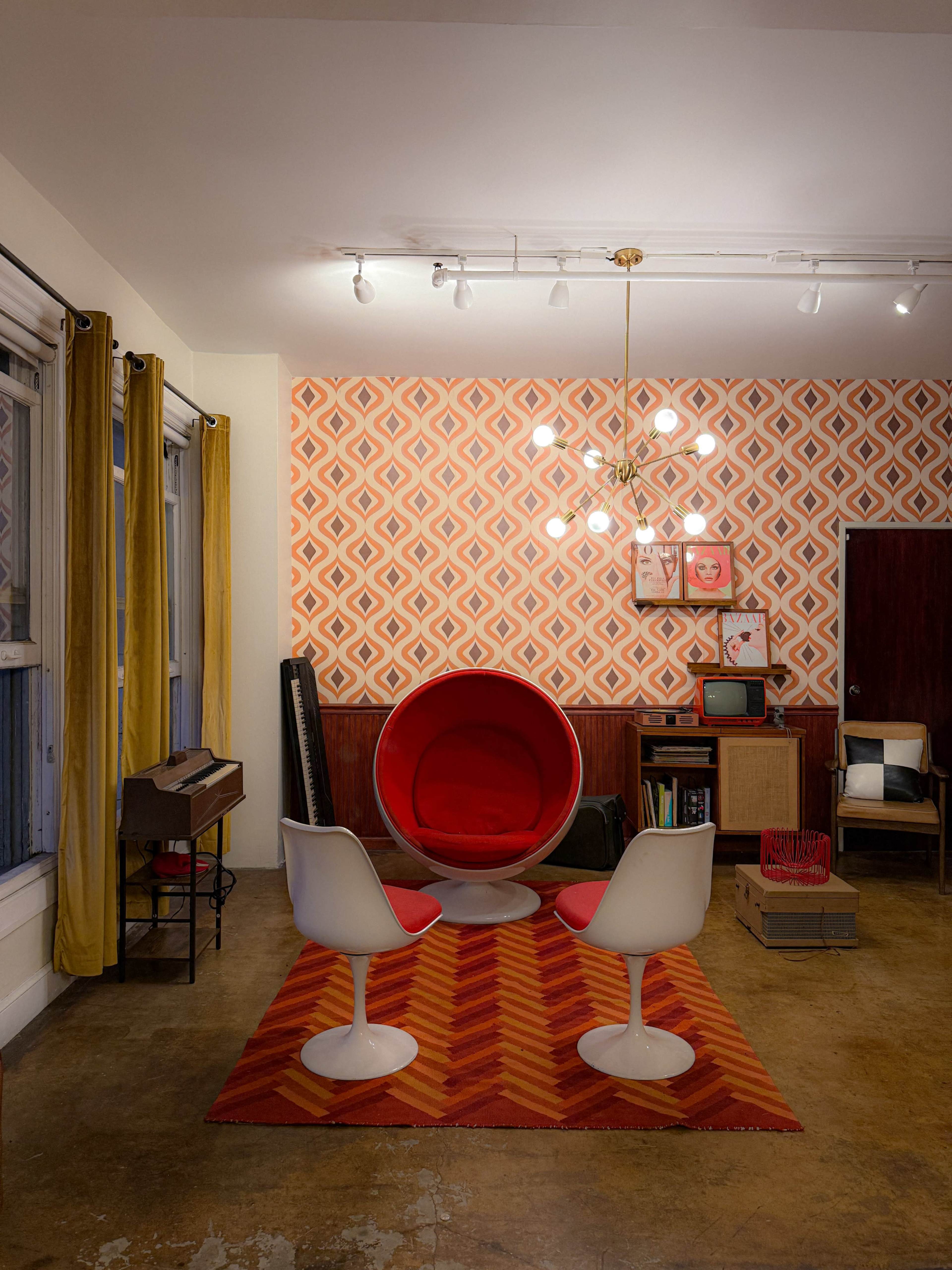The room features a red Egg chair, white tulip chairs, and a patterned orange wallpaper, complemented by a mid-century modern light fixture and wooden furnishings.