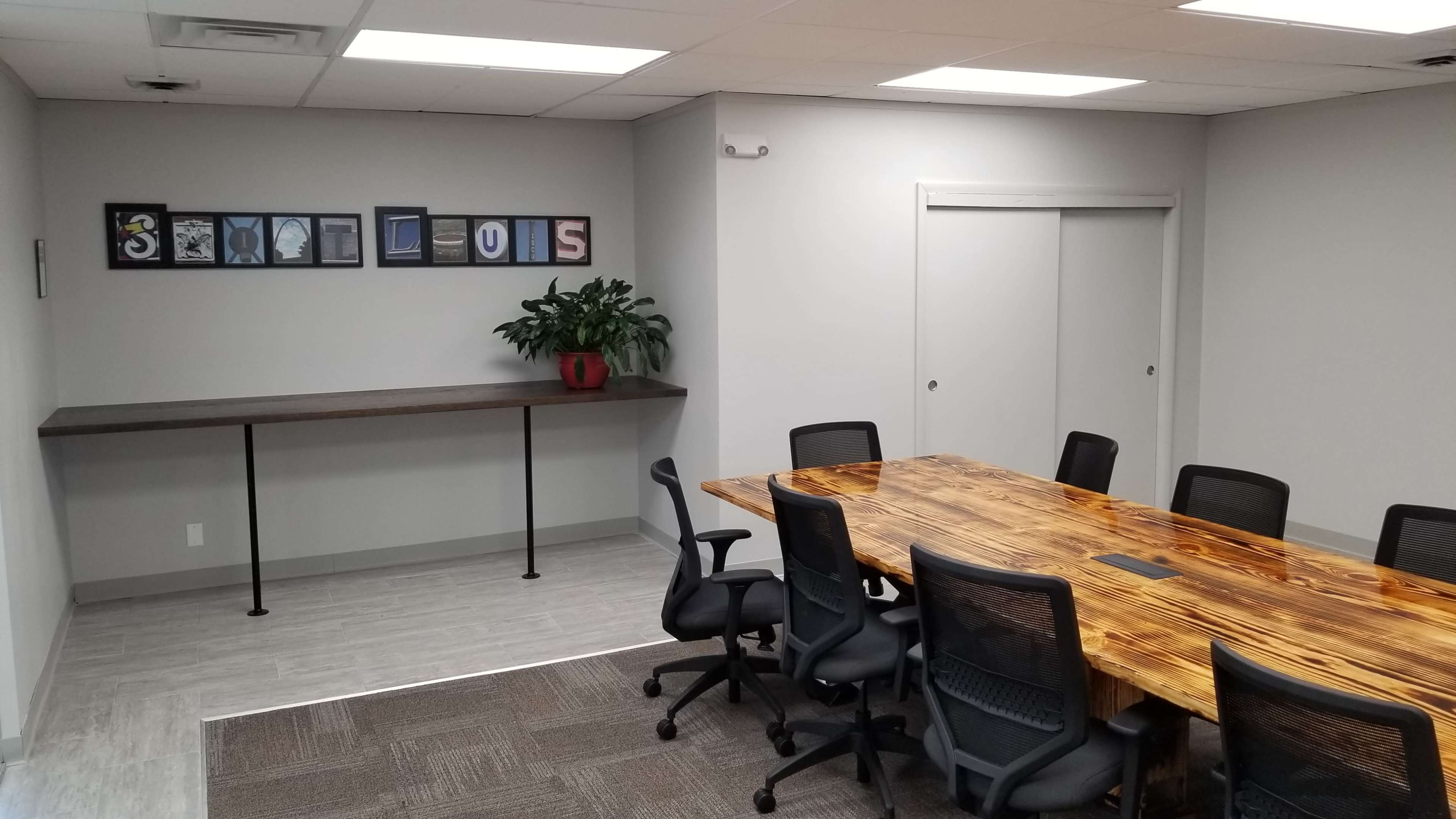 A conference room with a wooden table surrounded by black chairs, paired with a side shelf displaying framed artwork and a potted plant.