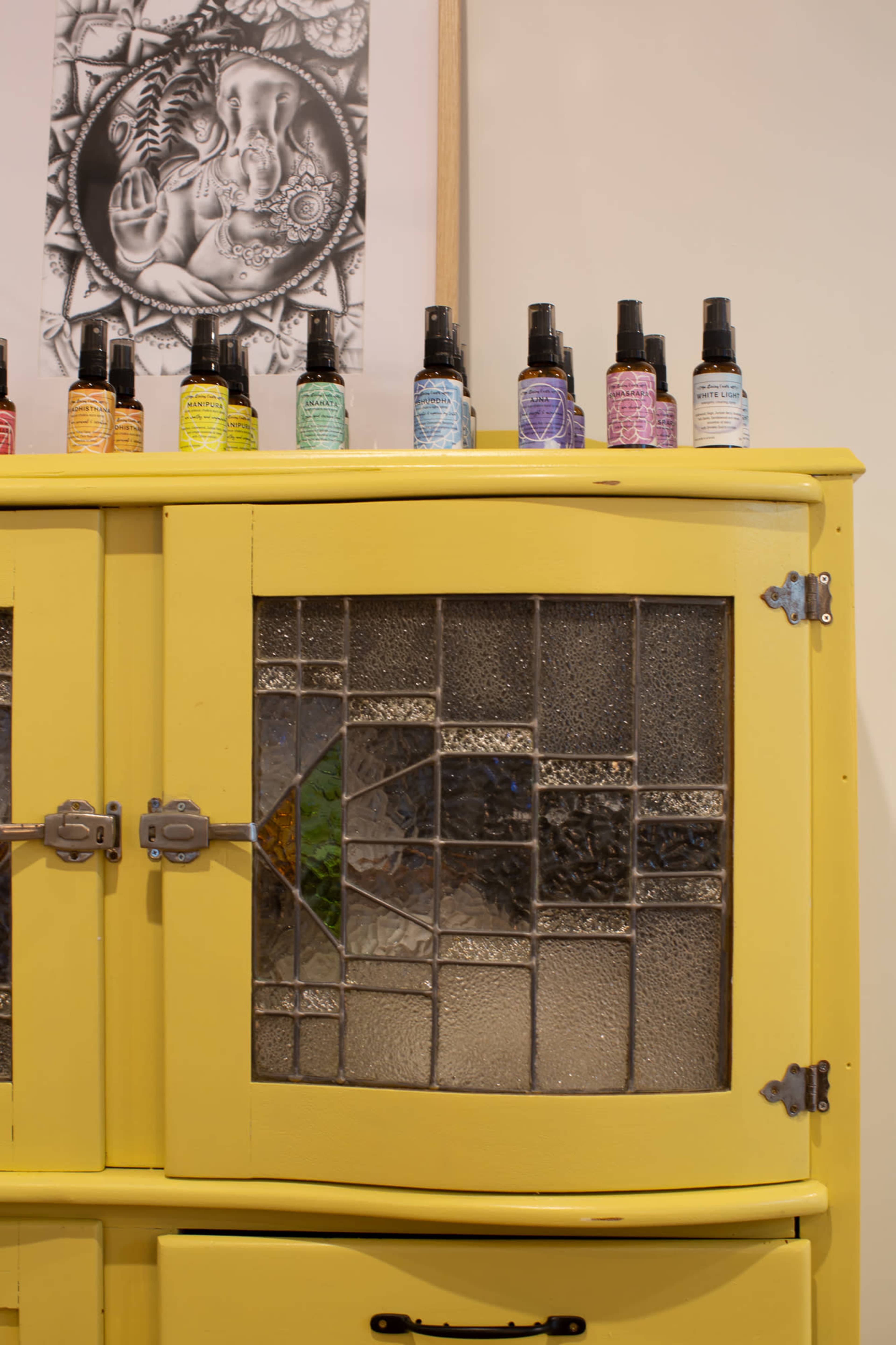 A yellow cabinet featuring a glass-paneled door displays various colored spray bottles on top and an artistic print of a figure behind it.