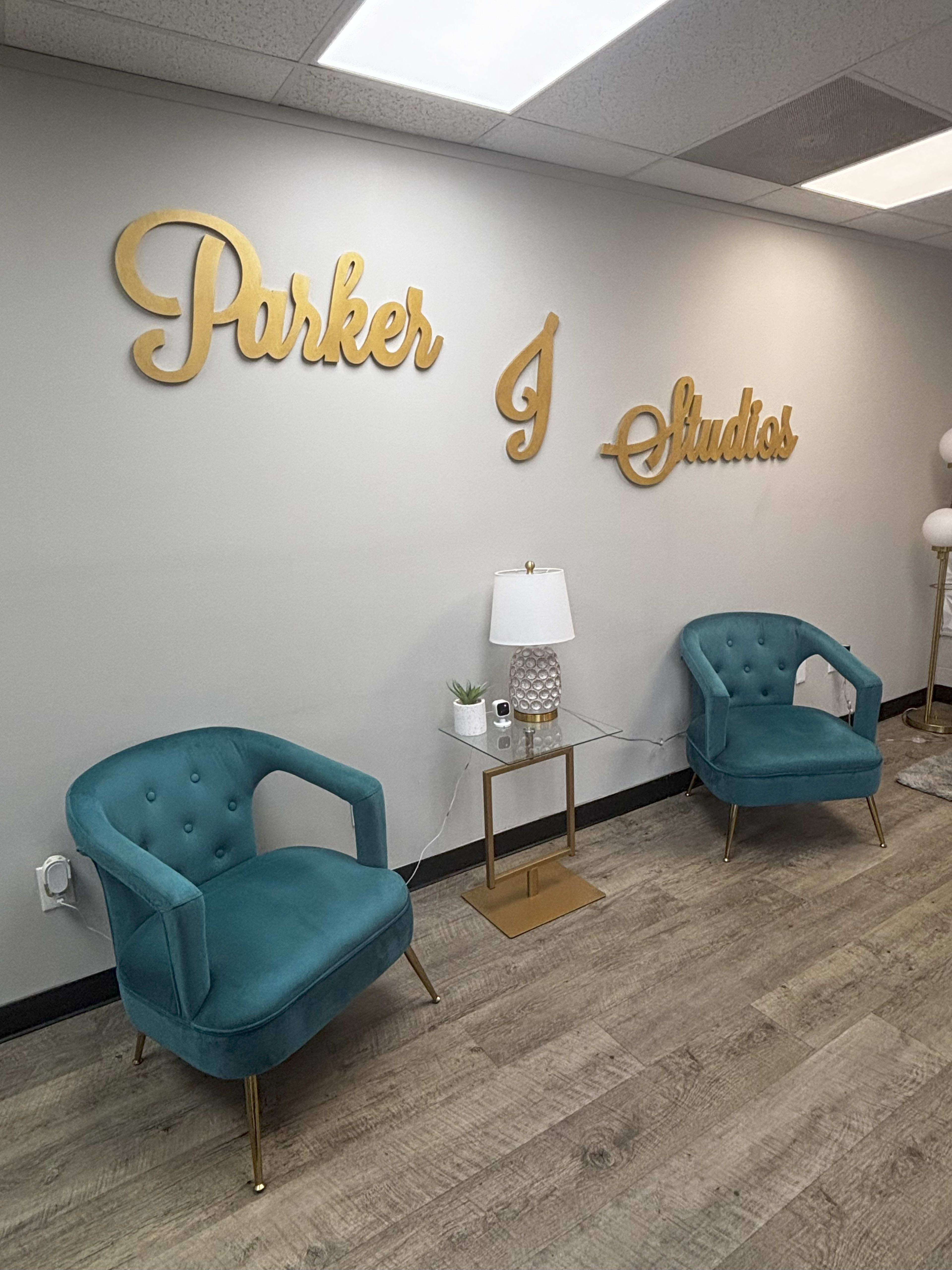 The image shows a waiting area with two teal chairs, a small gold side table holding a lamp, and a decorative plant against a wall featuring the words "Parker J Studios" in gold lettering.