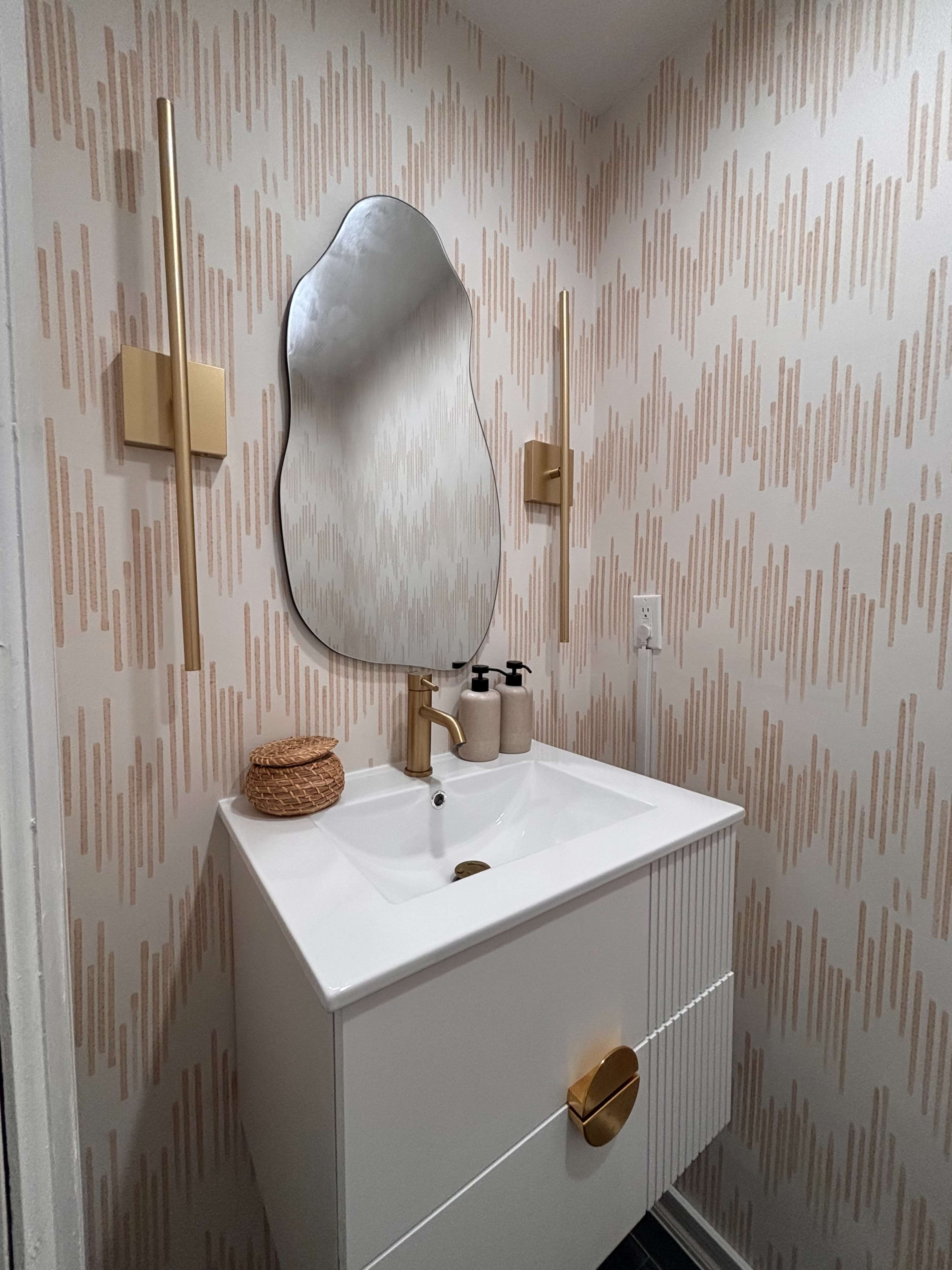 The image shows a modern bathroom with a stylish sink, a decorative mirror, and two gold wall sconces against a patterned wallpaper.