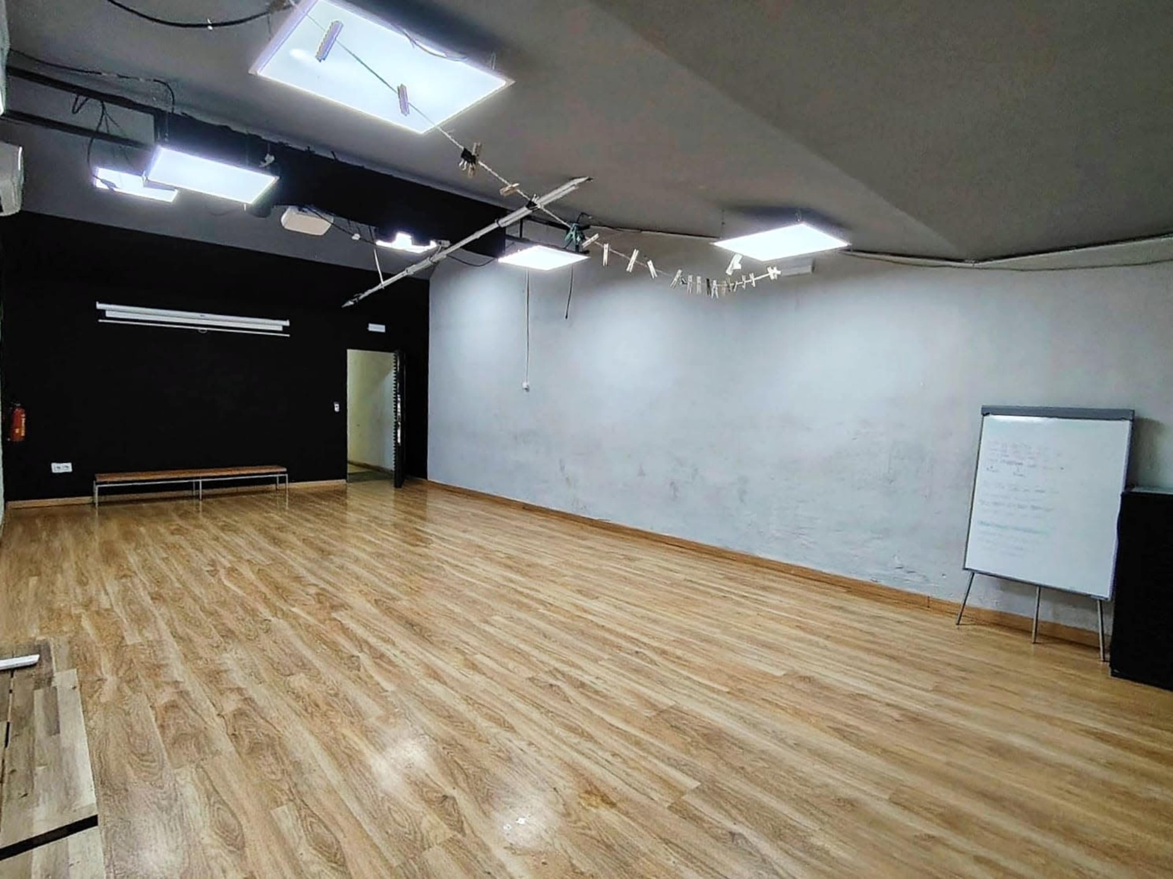 An empty room with wooden flooring, ceiling lights, and a whiteboard against one wall.
