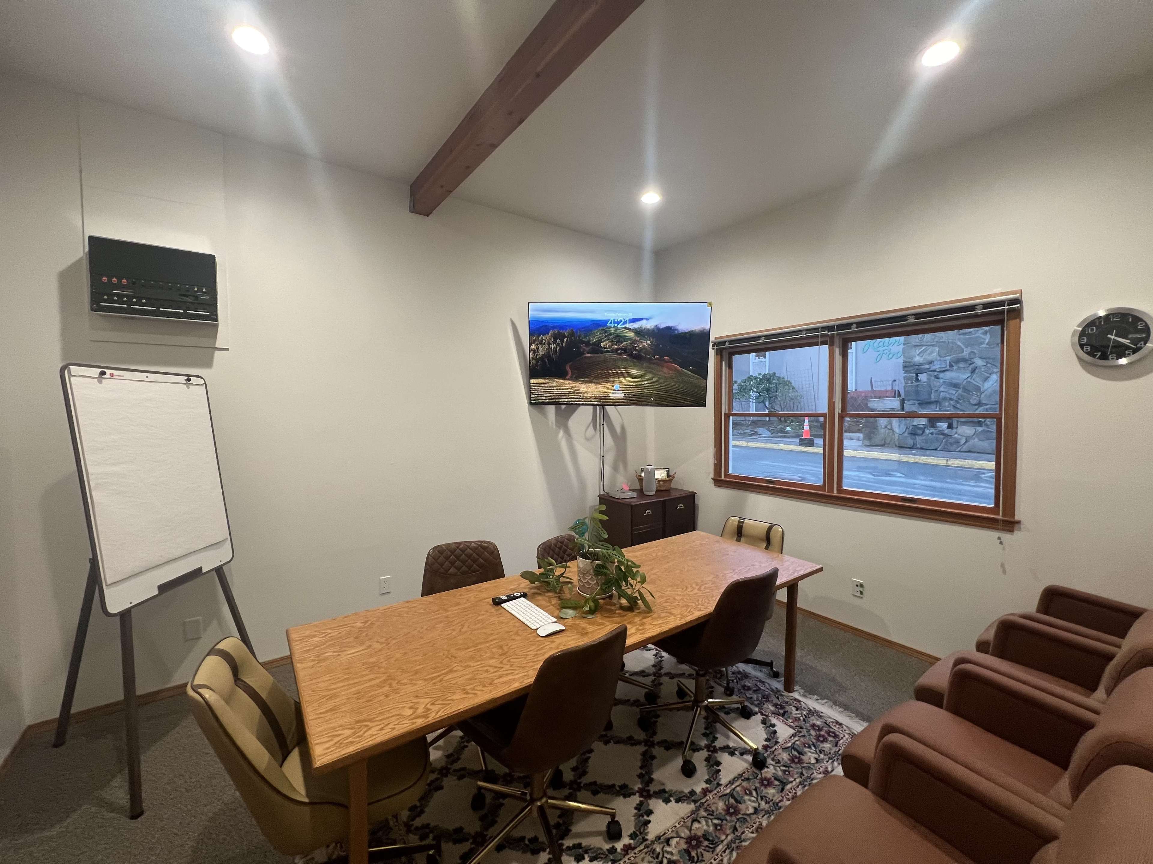 A small conference room features a wooden table surrounded by chairs, a television mounted on the wall, a whiteboard on a stand, and windows allowing natural light to enter.