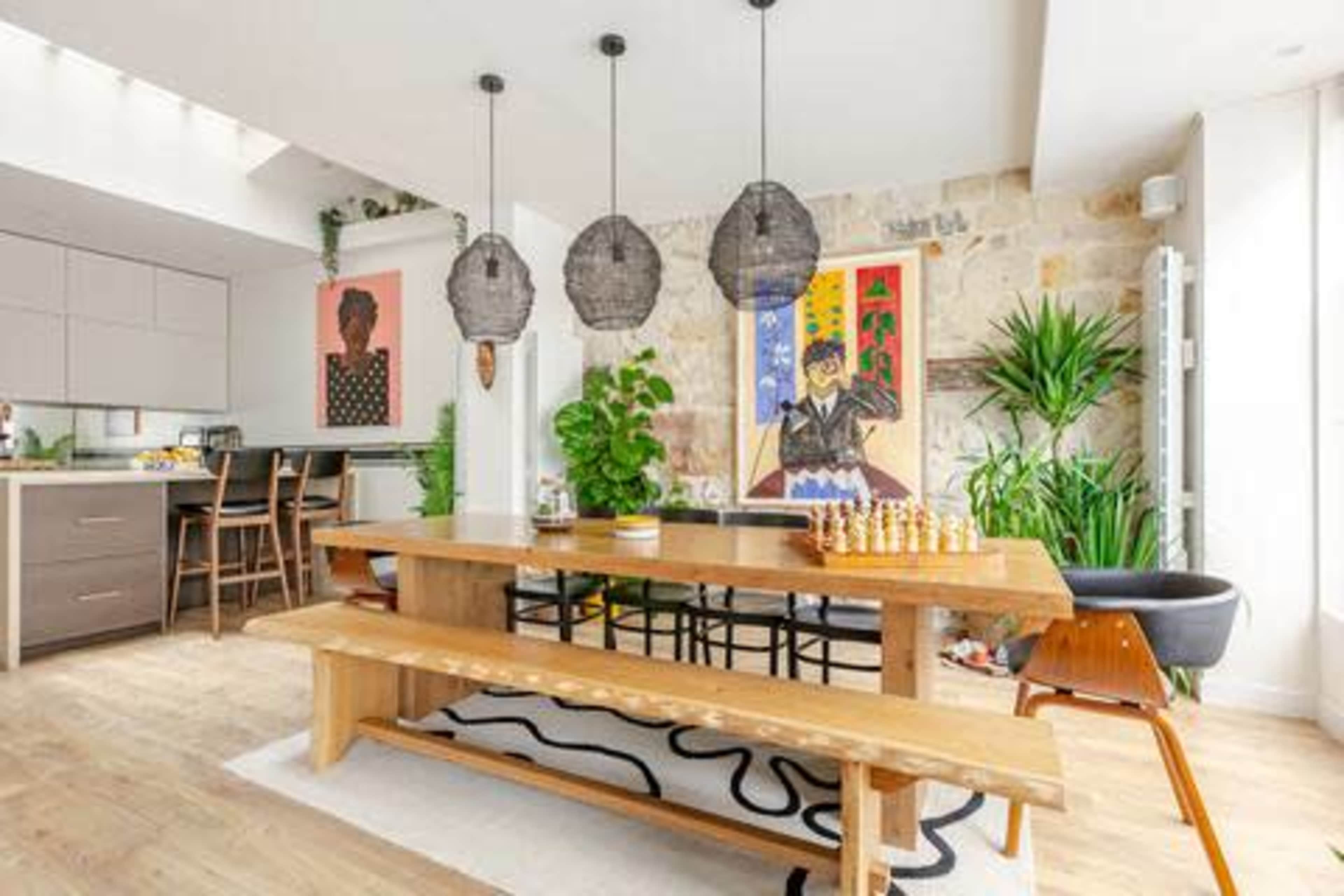 The image shows a modern dining area with a wooden table, bench seating, and hanging light fixtures, surrounded by plants and colorful artwork on the walls.