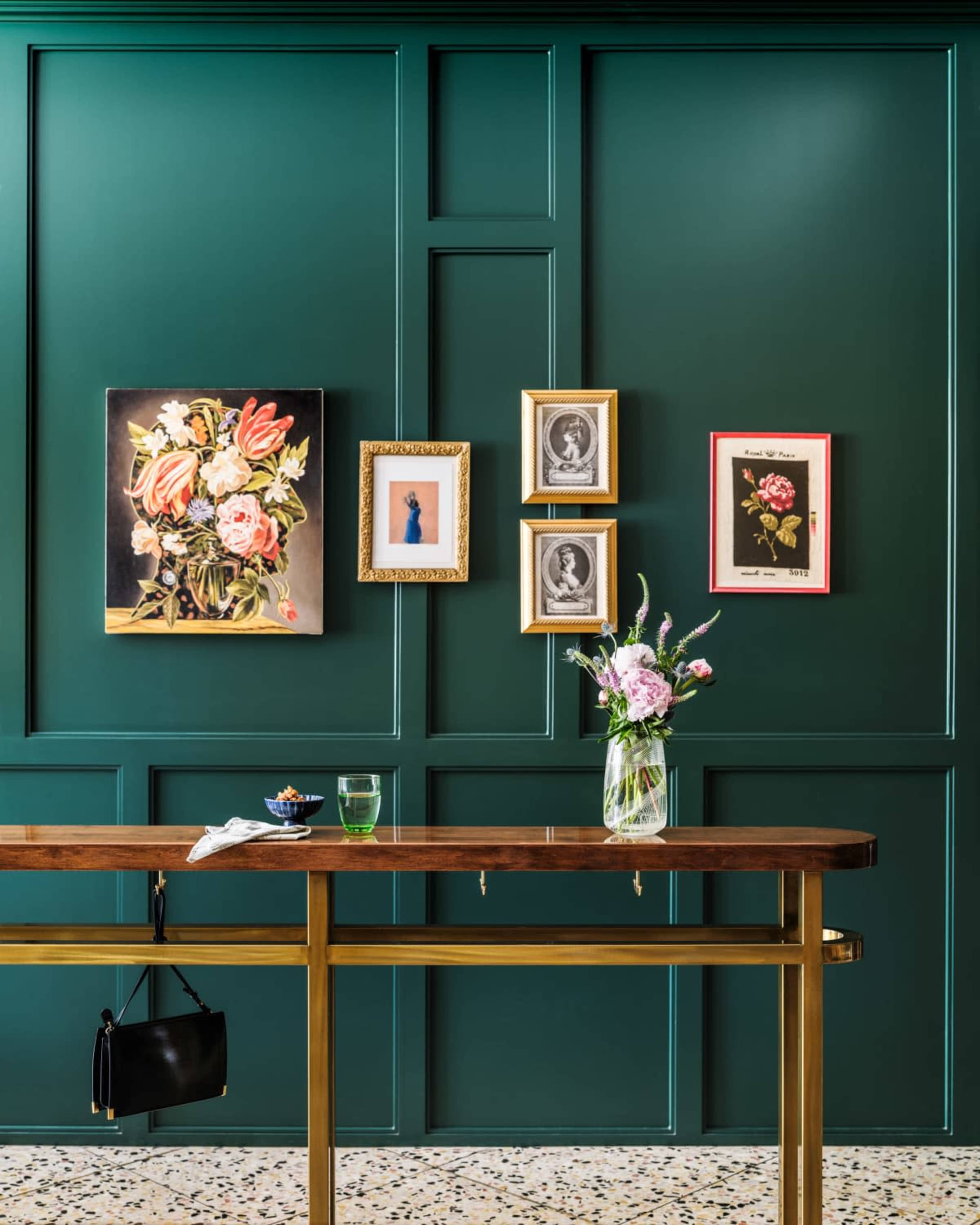 A dark green wall features a collection of framed artwork alongside a floral arrangement in a glass vase on a wooden table.