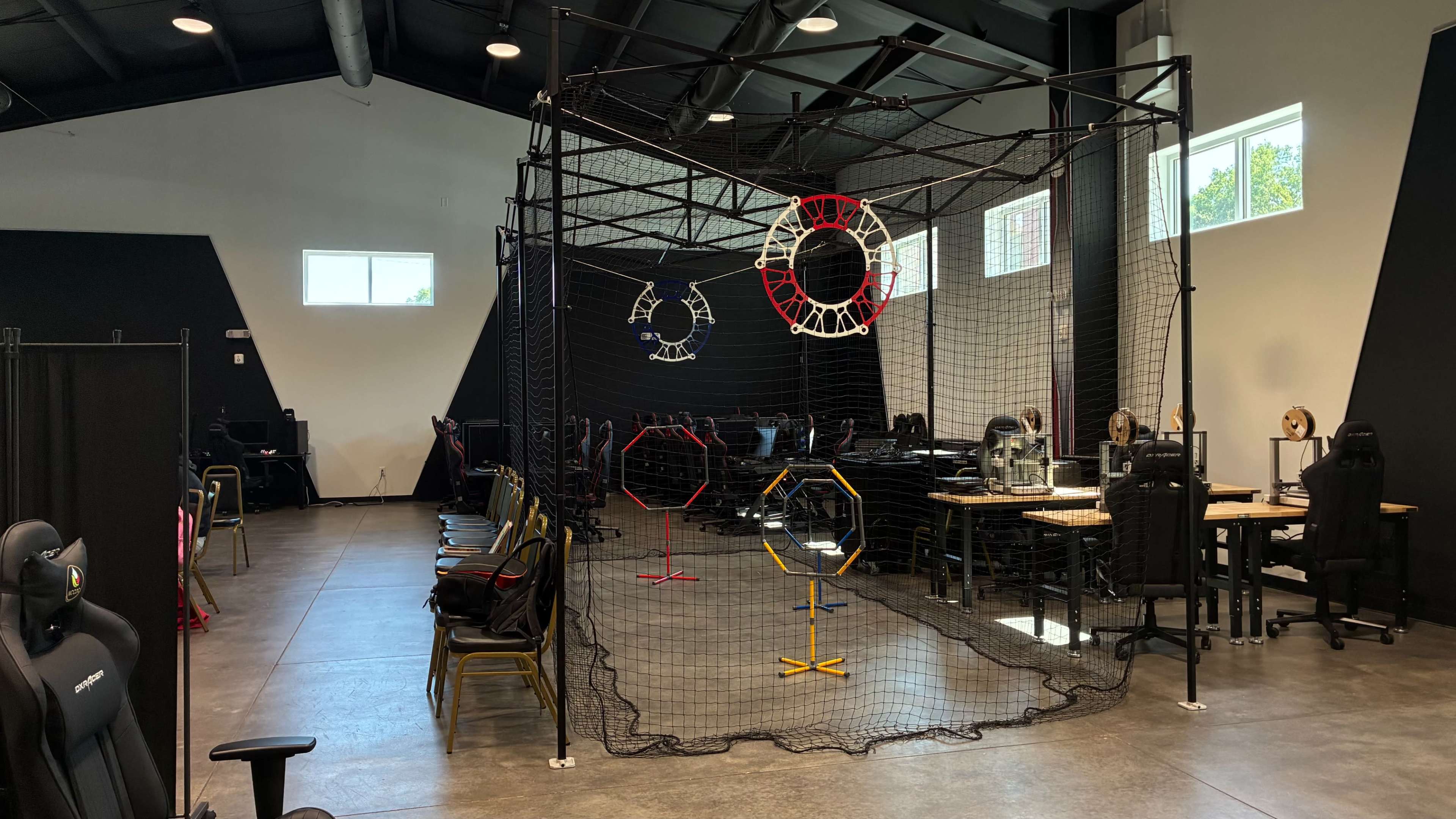 The image shows a spacious room with several computer desks, chairs, and a large net containing colorful hoop structures in the center.