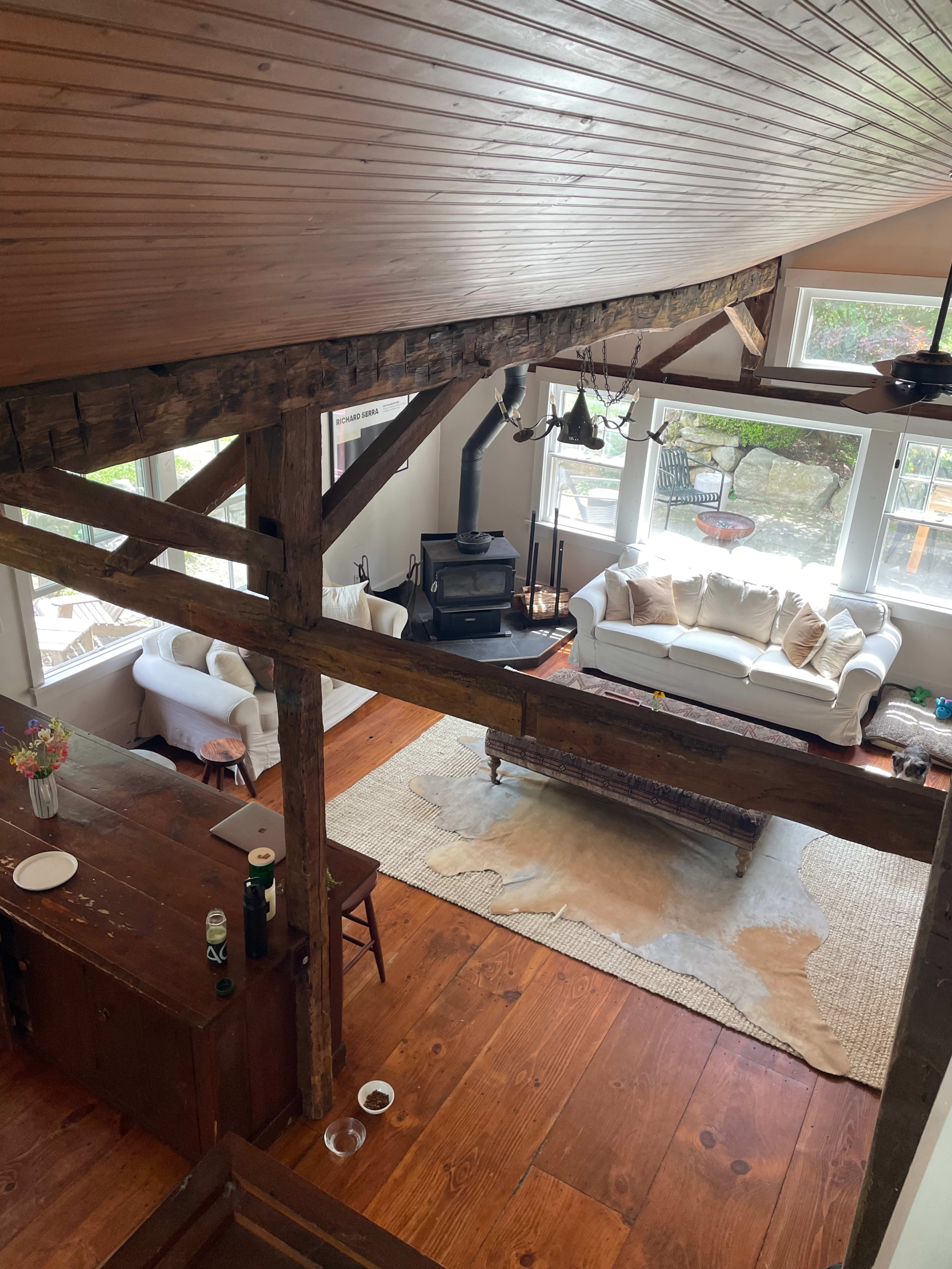 A spacious living area with wooden beams, a white couch, a rustic coffee table on a rug, and large windows allowing natural light.