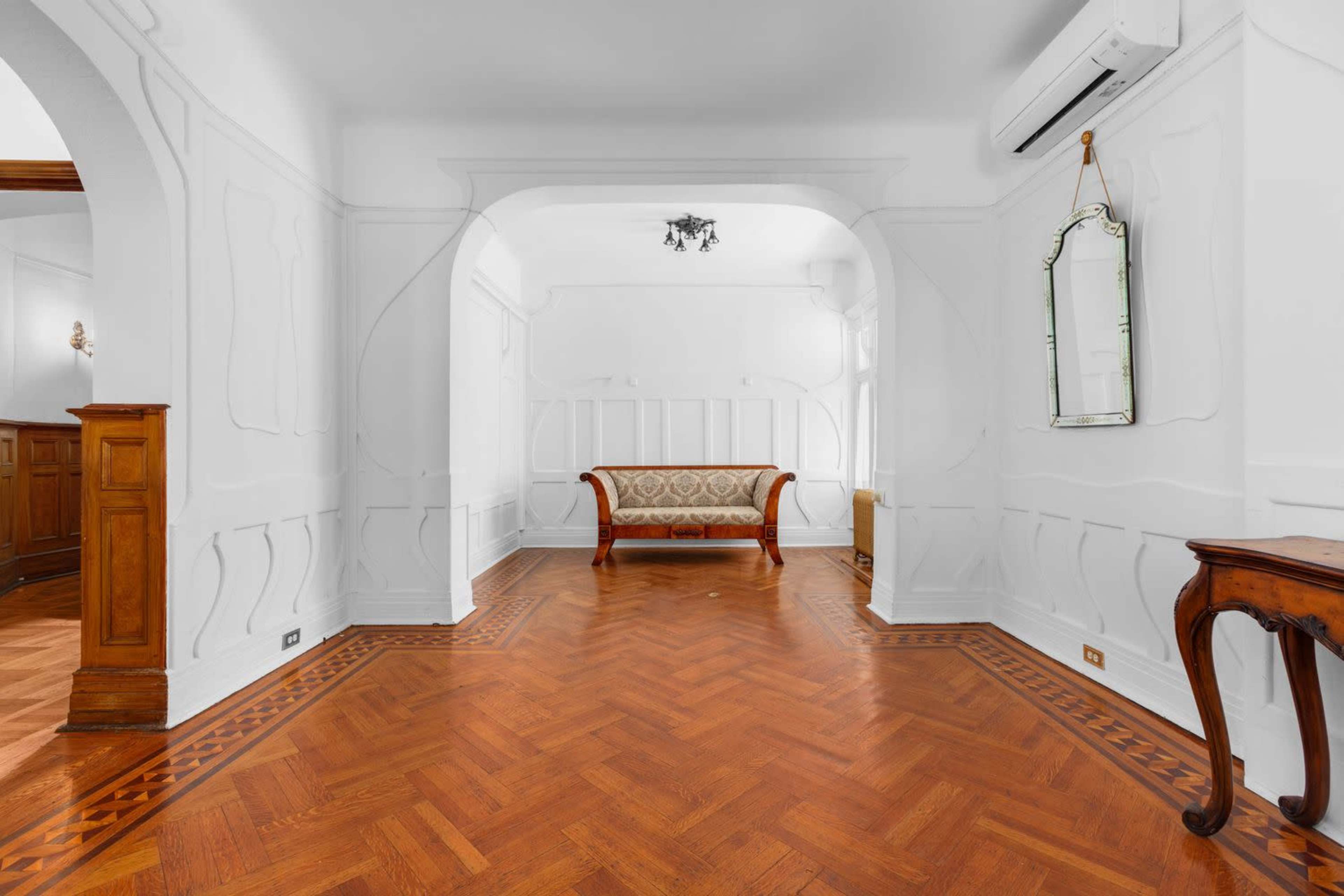 A spacious, empty room with wooden herringbone flooring and a decorative archway leading to another area, featuring a vintage sofa and a wall mirror.