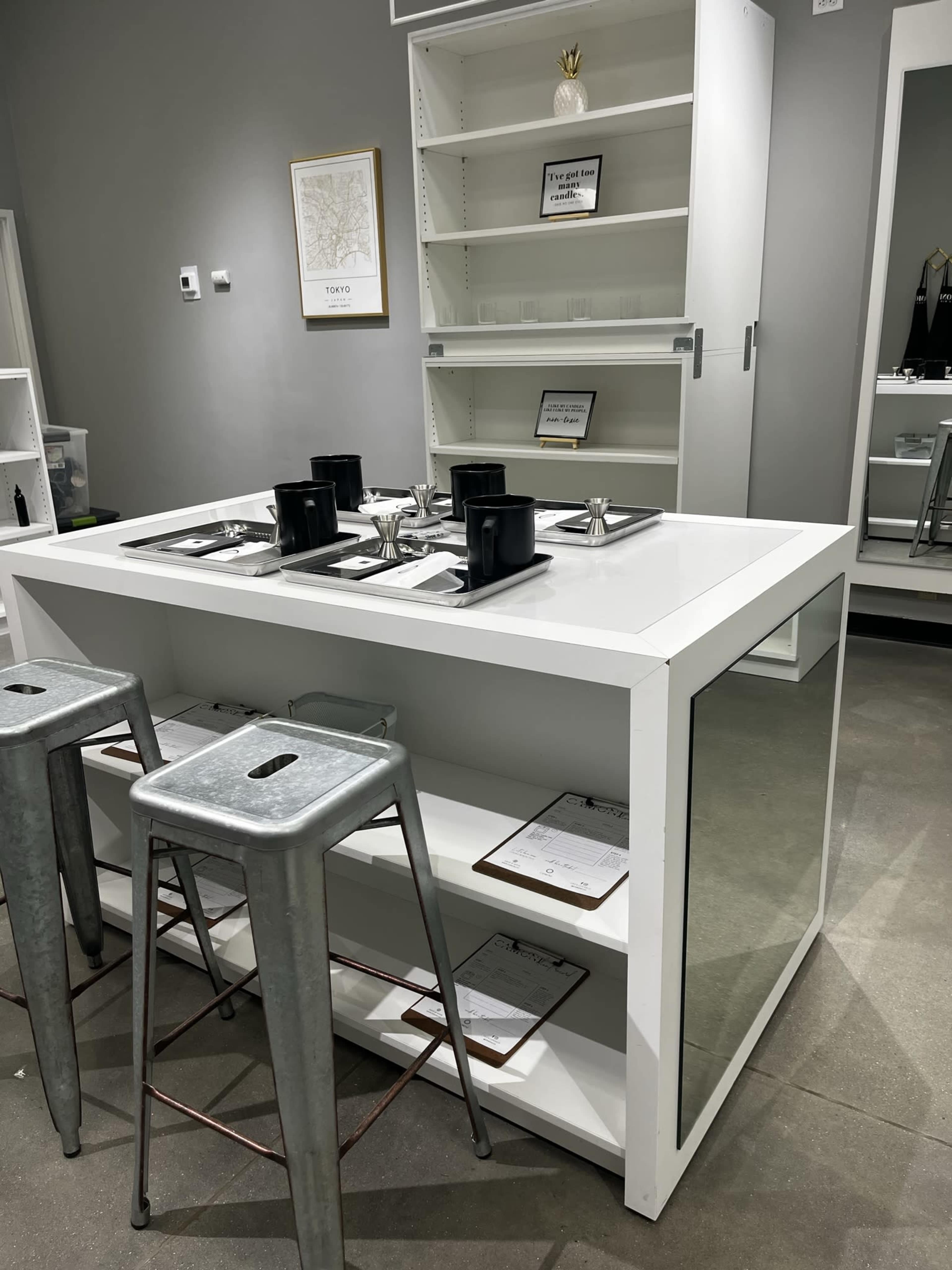 A modern white table with two metal stools, situated in a minimalist room with shelves and decor items in the background.
