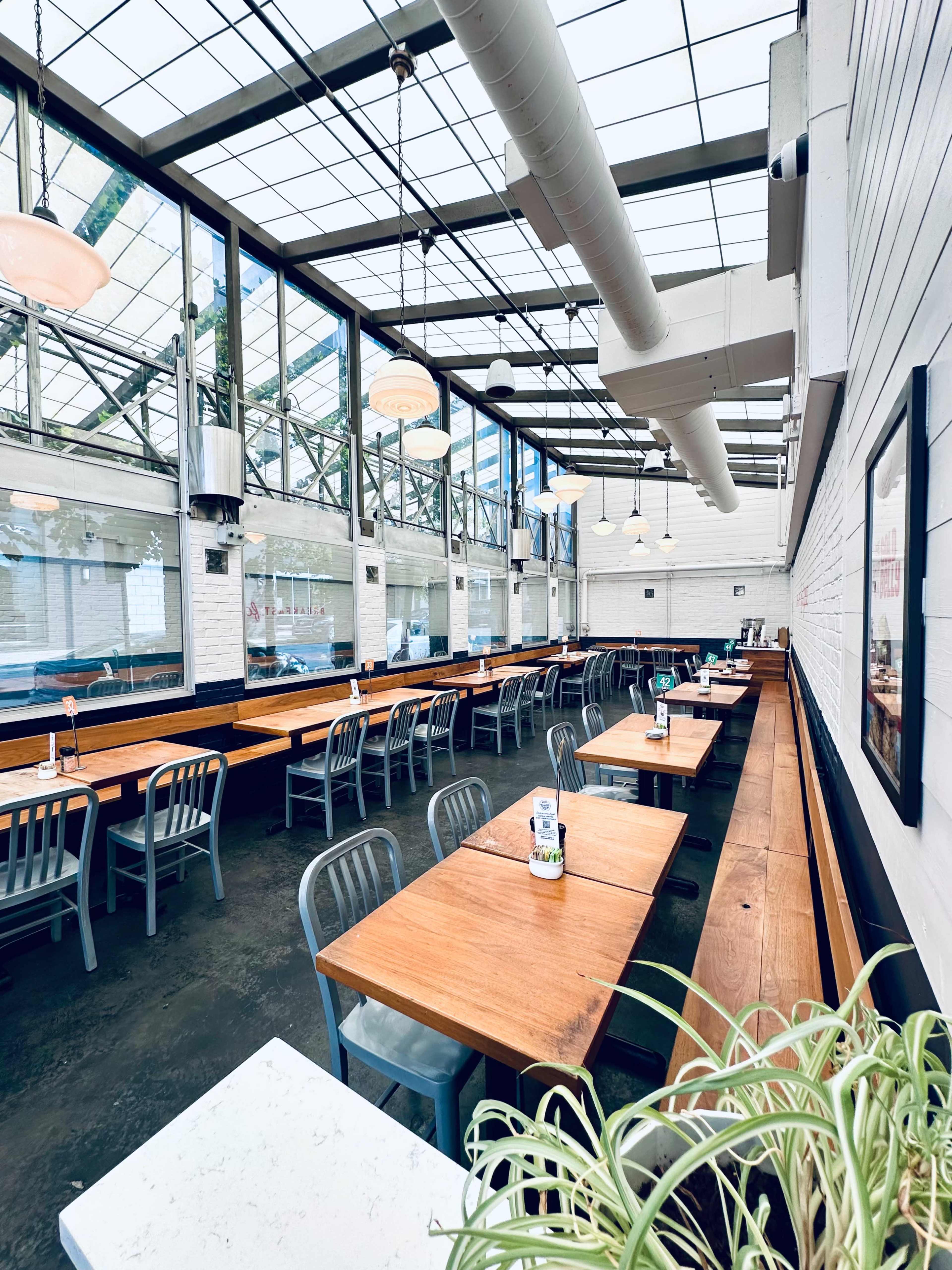 The image shows a bright, modern restaurant interior featuring rows of wooden tables and chairs under a glass roof.