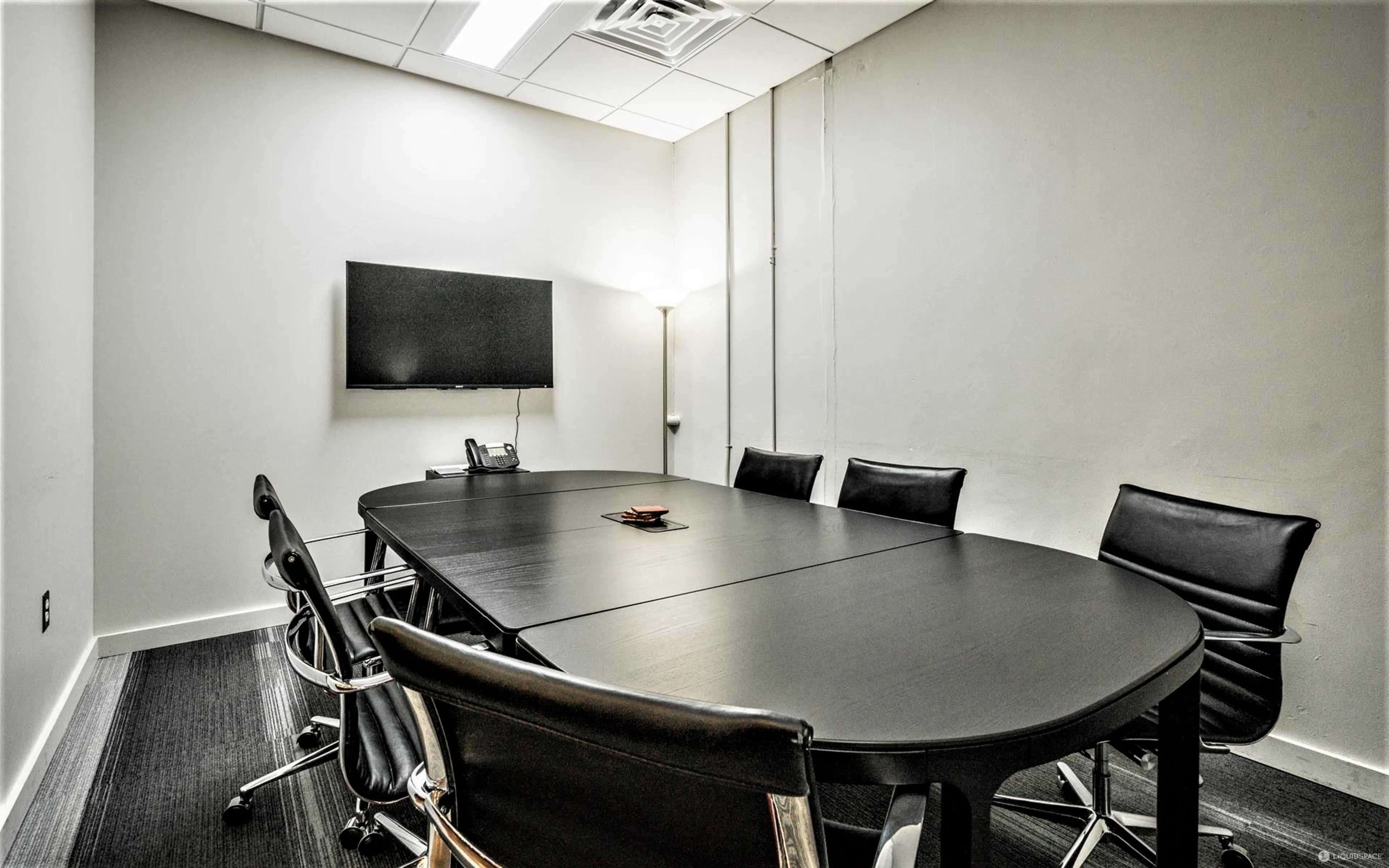 A small conference room features a large oval table surrounded by black leather chairs, with a wall-mounted TV and a lamp in the corner.