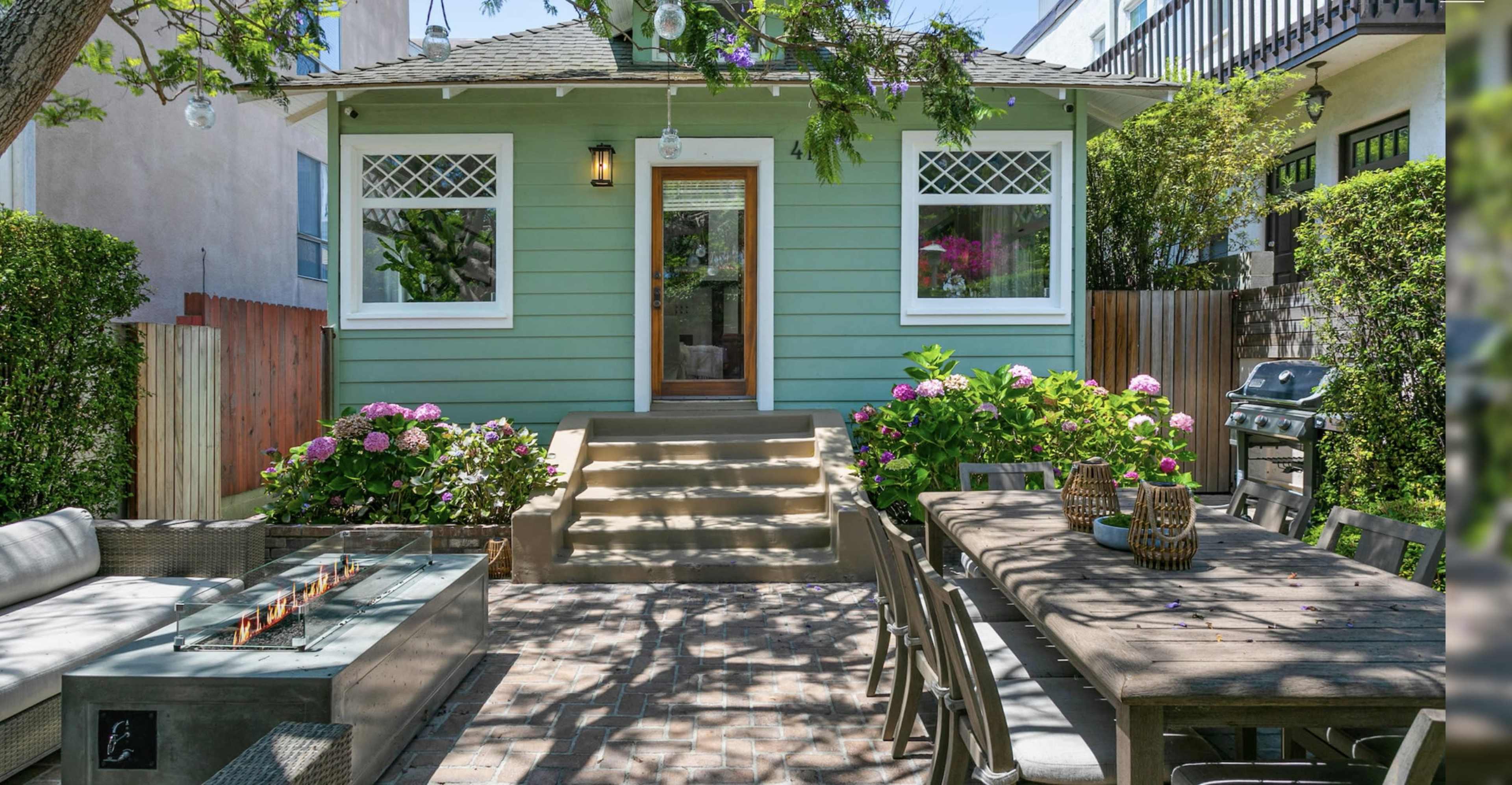 A light green house with a landscaped front yard features a staircase leading to a wooden door, with outdoor seating and a grill nearby.