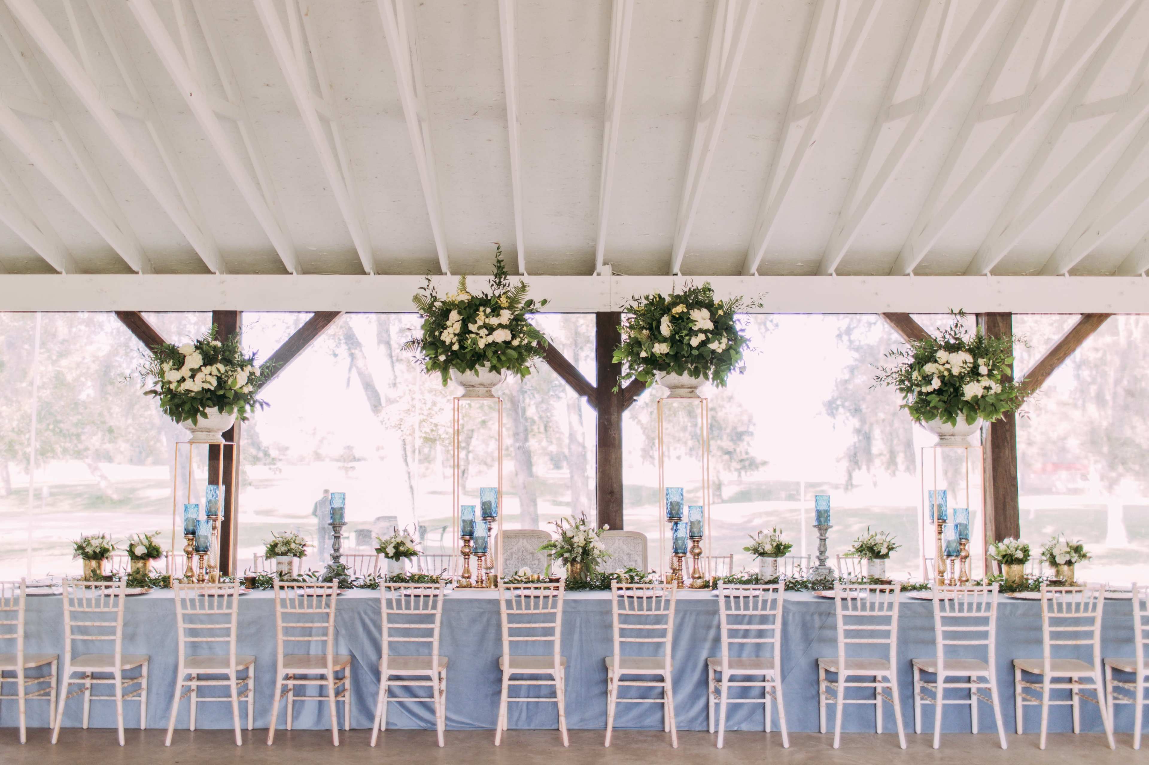 A long banquet table with floral centerpieces and blue table runners is set under a white-beamed pavilion.