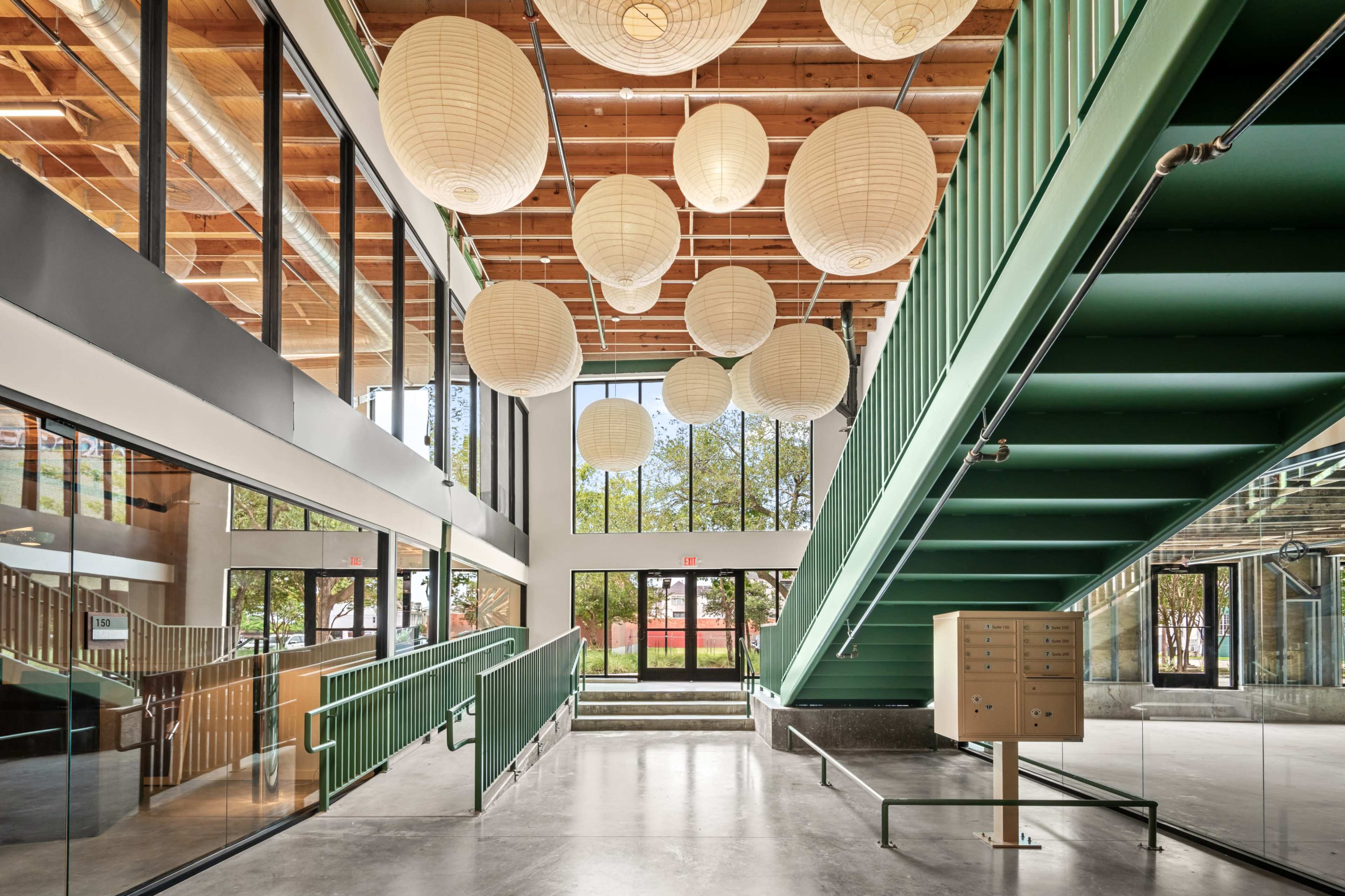 The image shows a modern, open interior space featuring a staircase with green railings, large windows, and round paper lanterns hanging from the ceiling.