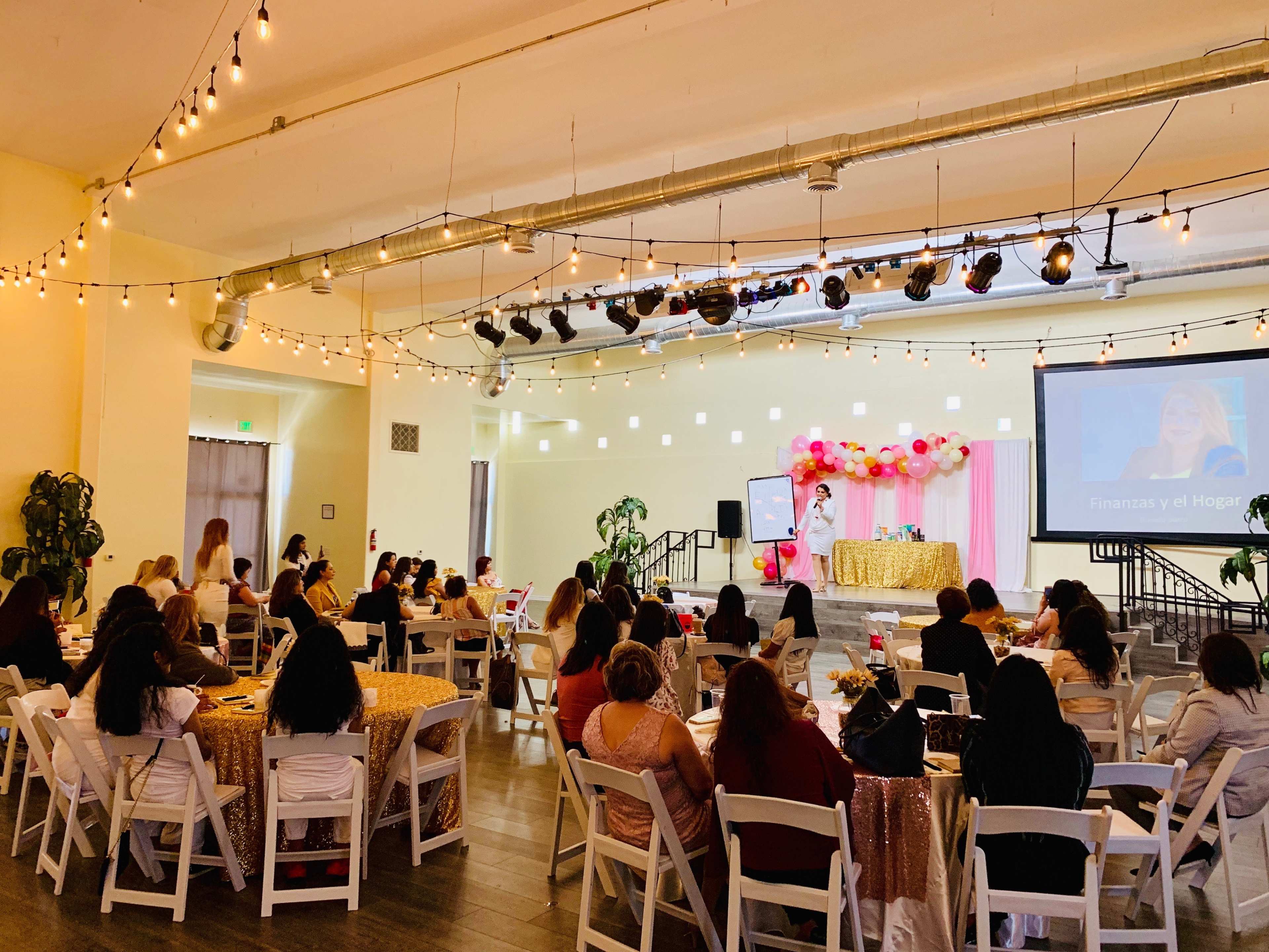 A group of attendees sits at tables in a spacious venue decorated with balloons and lights, while a speaker presents on stage.