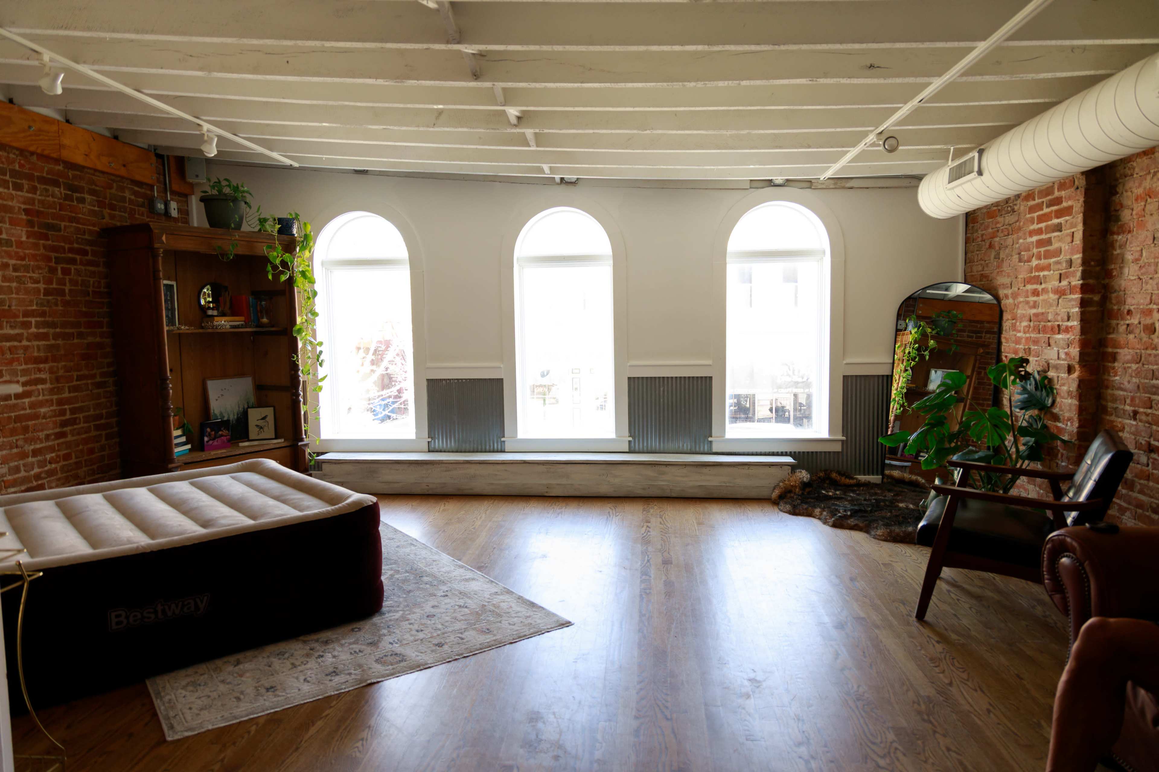 The image shows a spacious room with brick walls, featuring a large window with three arches, an inflatable mattress, a bookshelf, plants, and a chair.