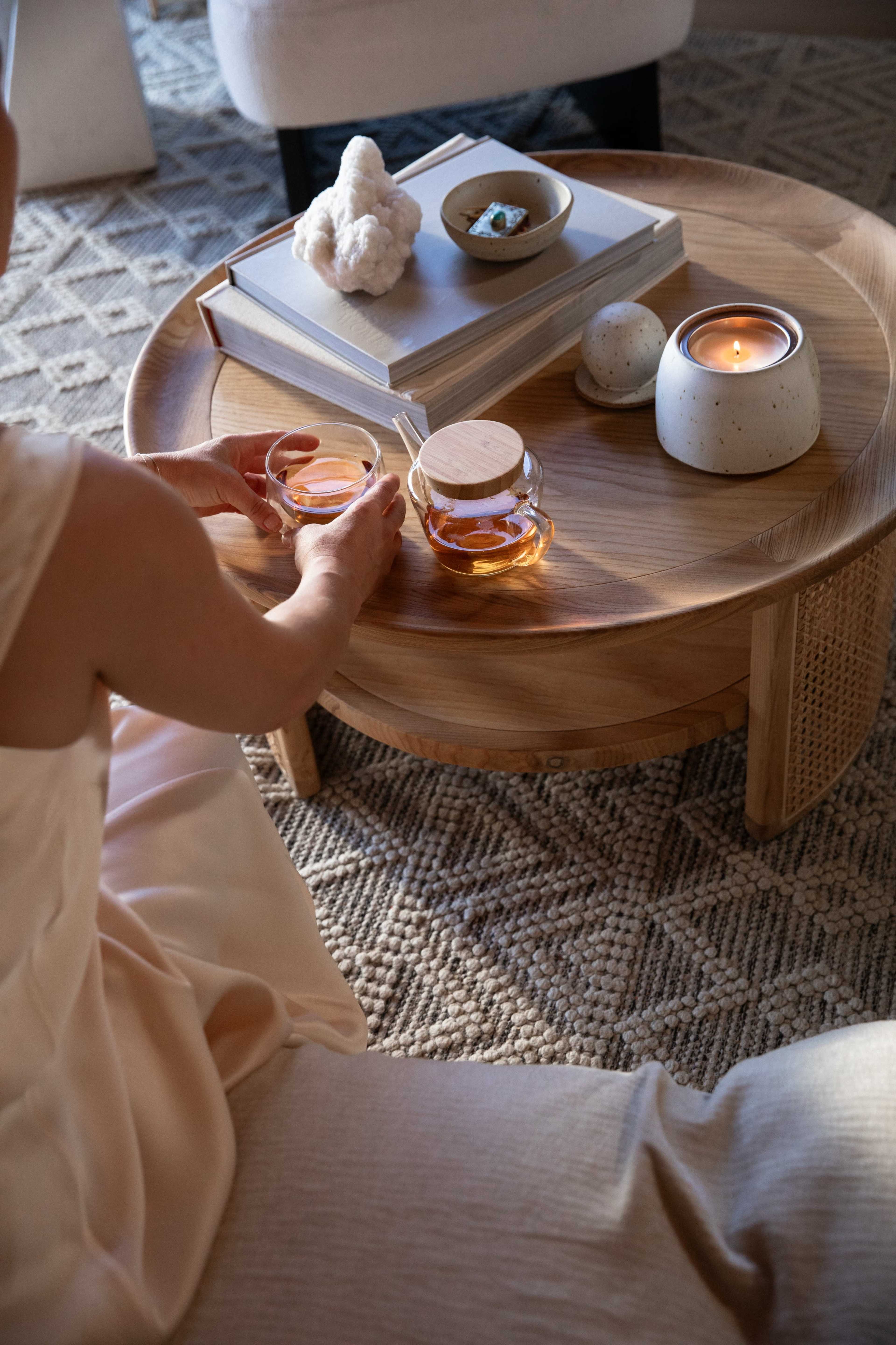 A person sits on a cushion, holding two glasses of tea while positioned near a low wooden coffee table decorated with a book, a bowl of food, and a candle.