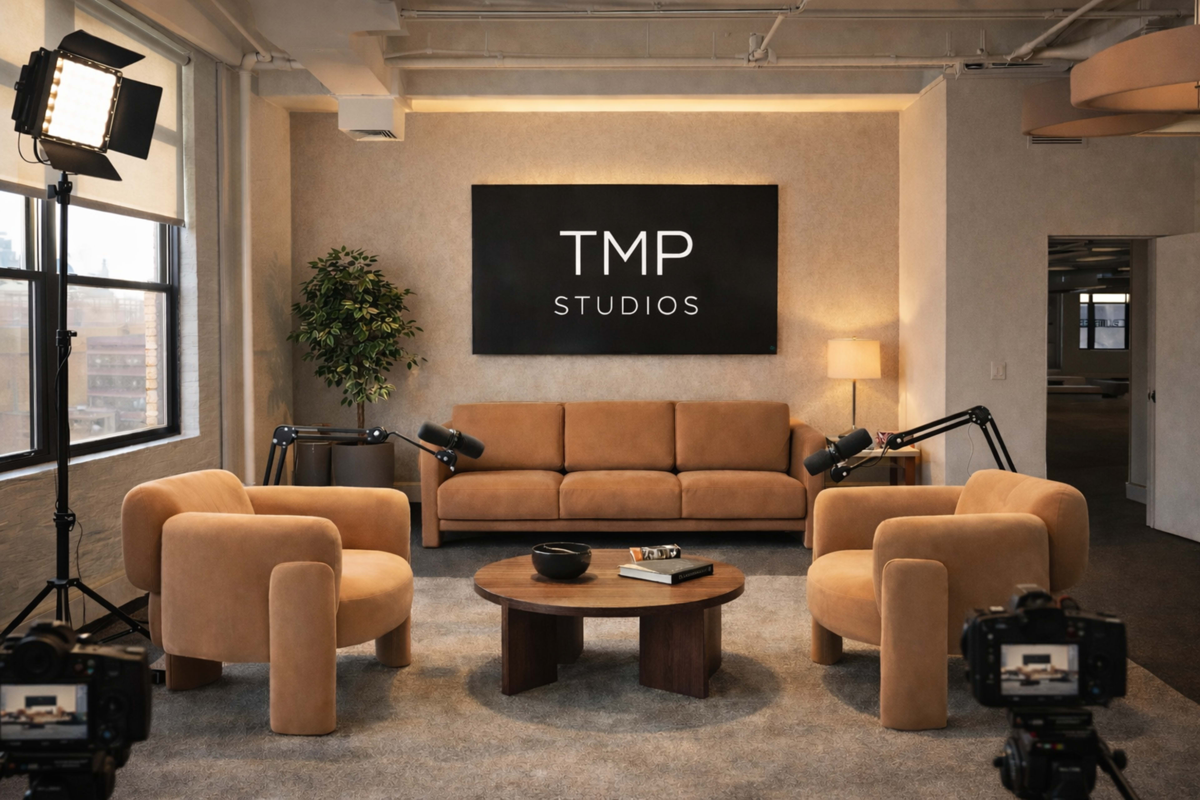 A modern studio interior features a brown couch and two matching armchairs, with a wooden table in the center and a large black sign that reads "TMP STUDIOS" on the wall.