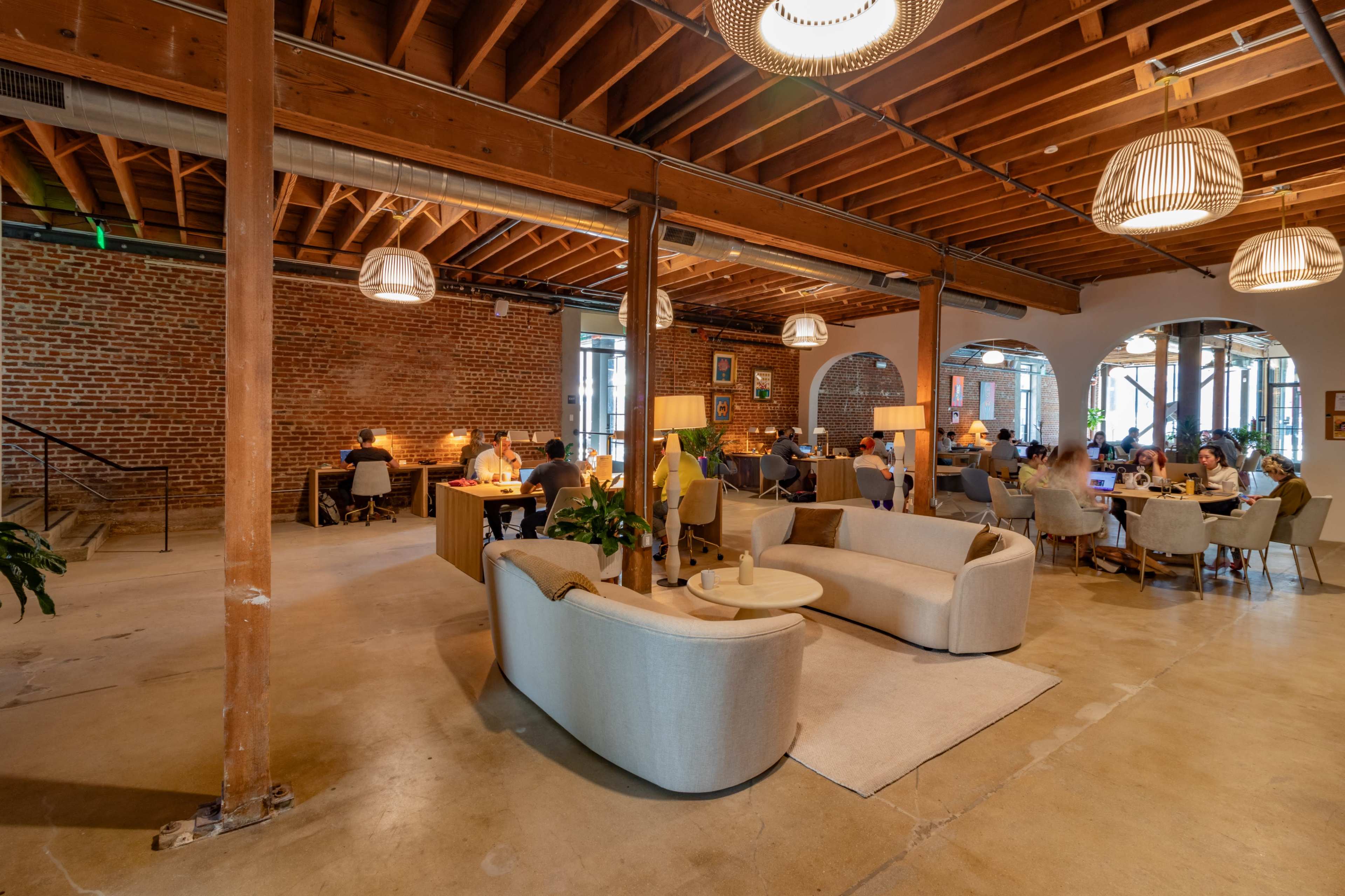A spacious, modern café with wooden beams, brick walls, and comfortable seating areas filled with patrons engaged in conversation or working.