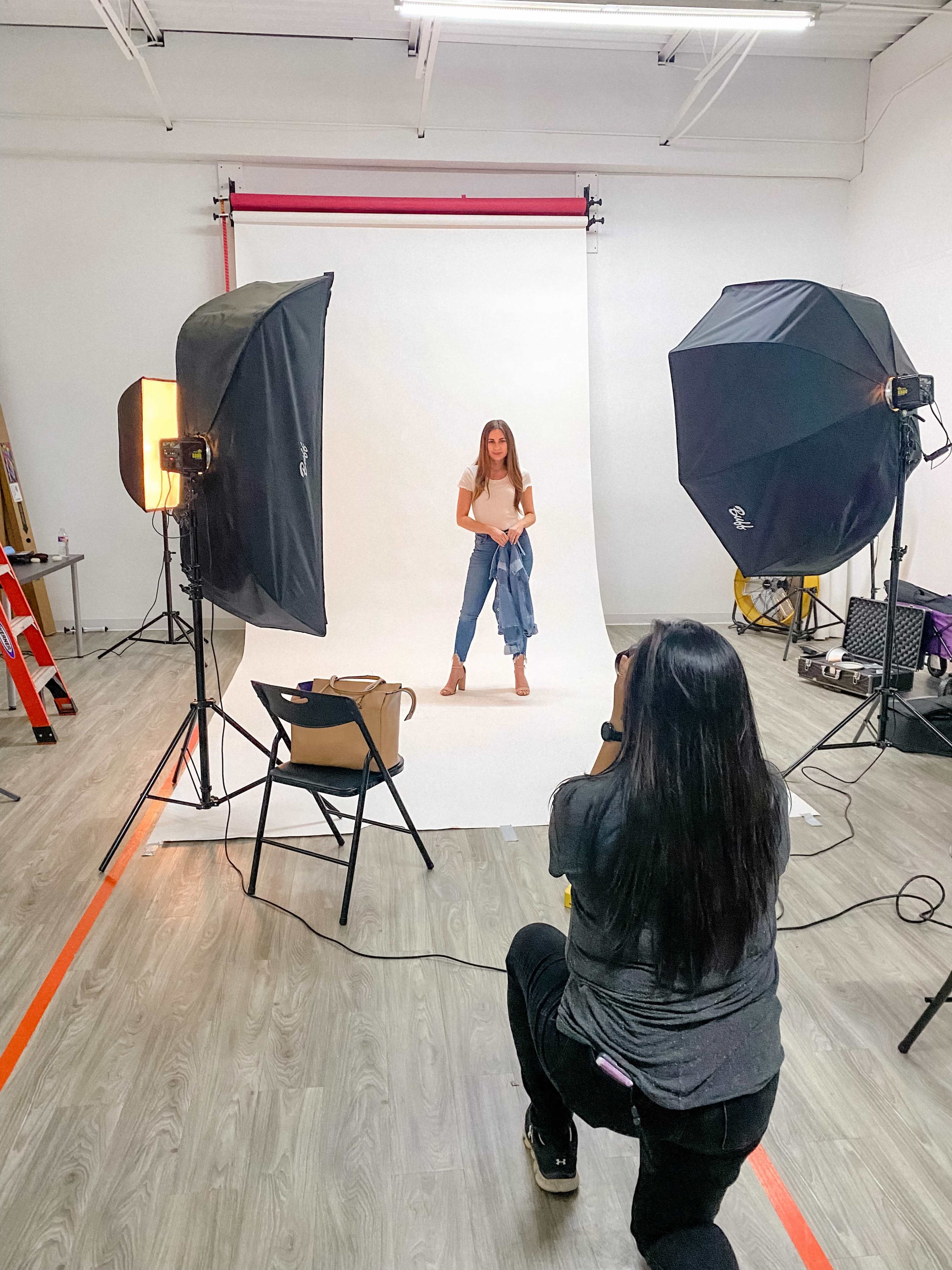 A photographer is capturing an image of a model posing in front of a backdrop in a studio equipped with lighting setups.