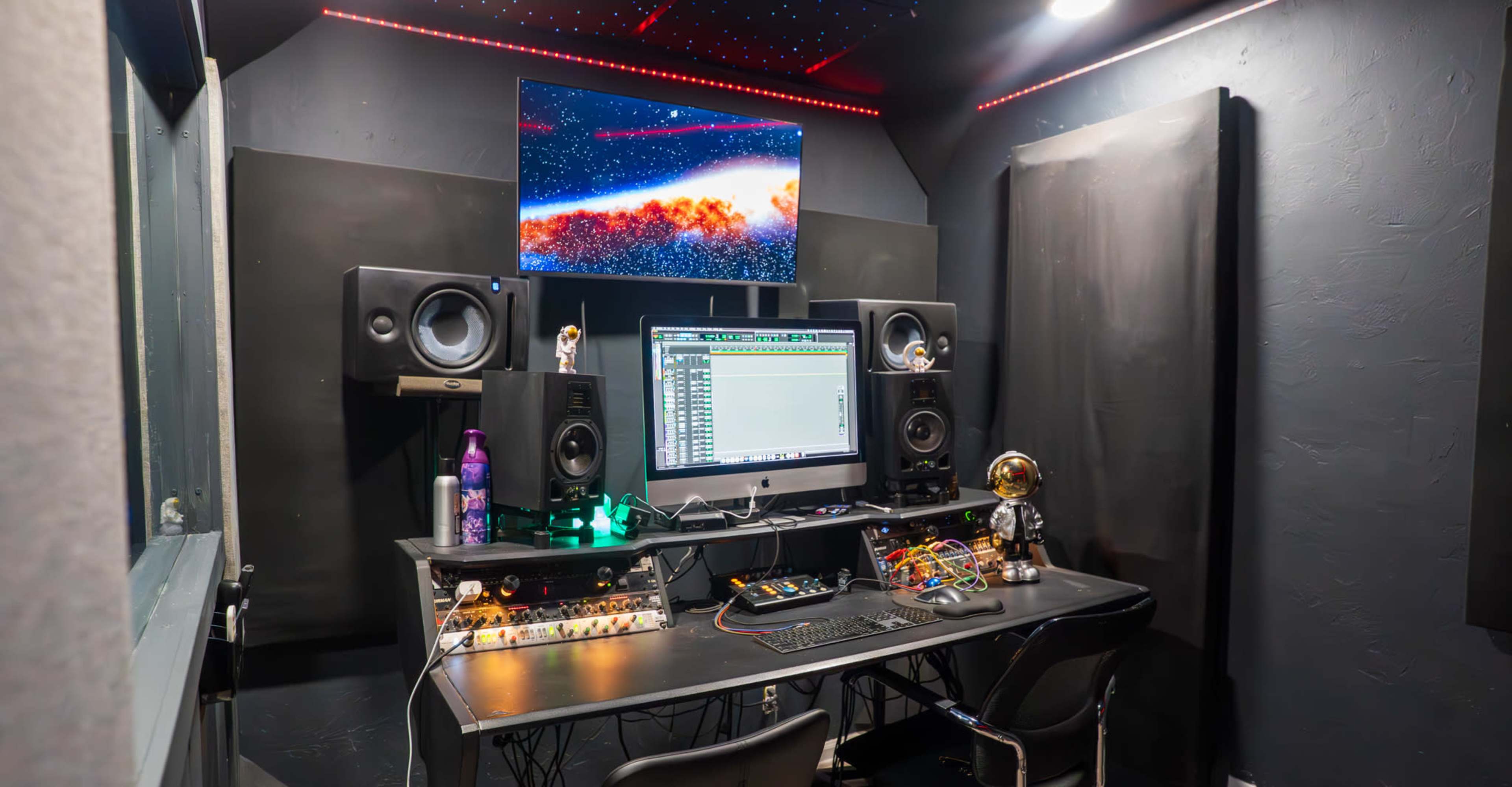 The image shows a music studio with a computer monitor displaying audio software, speakers on either side, and various equipment on a black desk, all under ambient lighting.