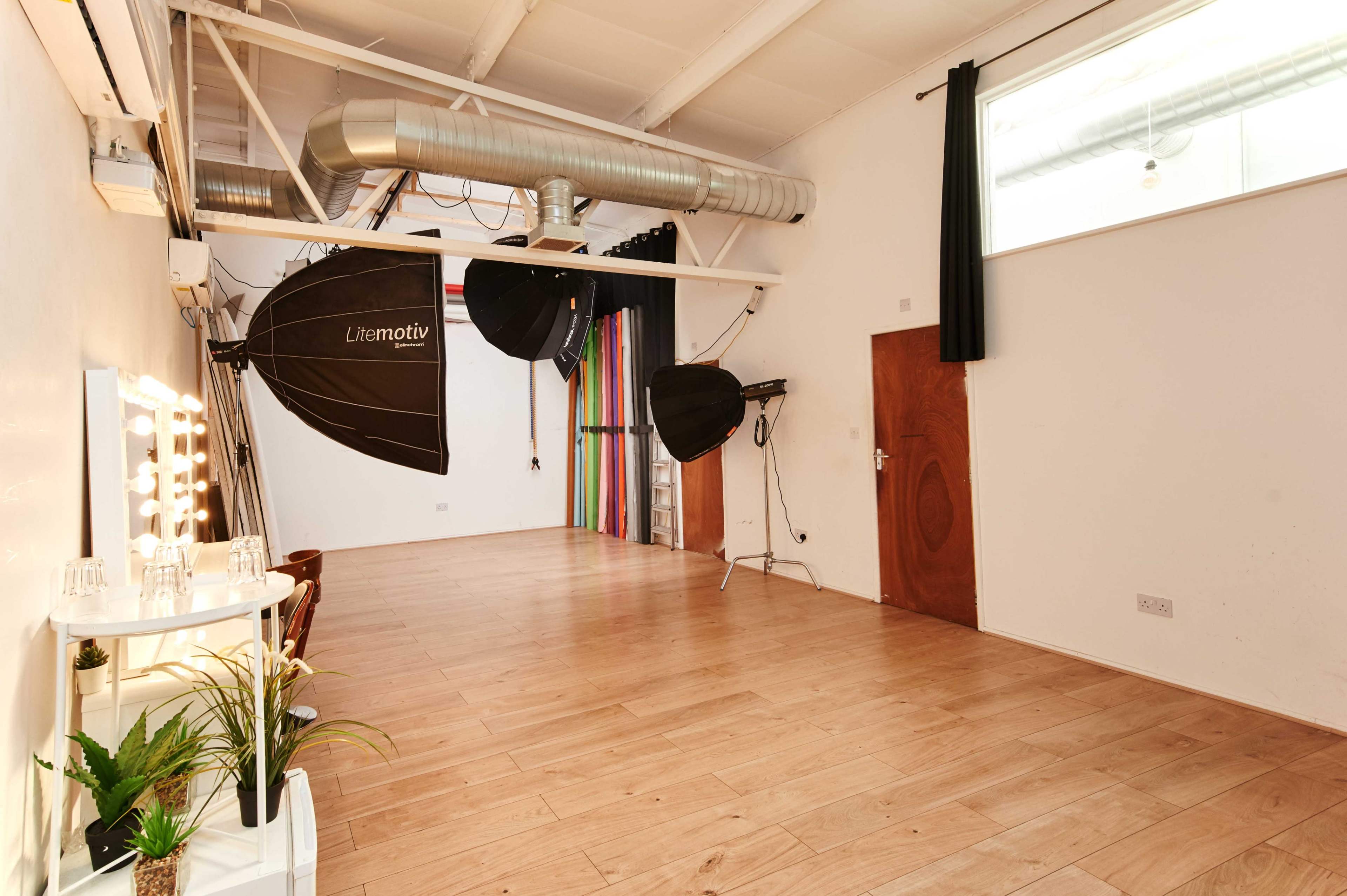 A spacious studio with wooden flooring, equipped with large softbox lights and a mirrored vanity station.