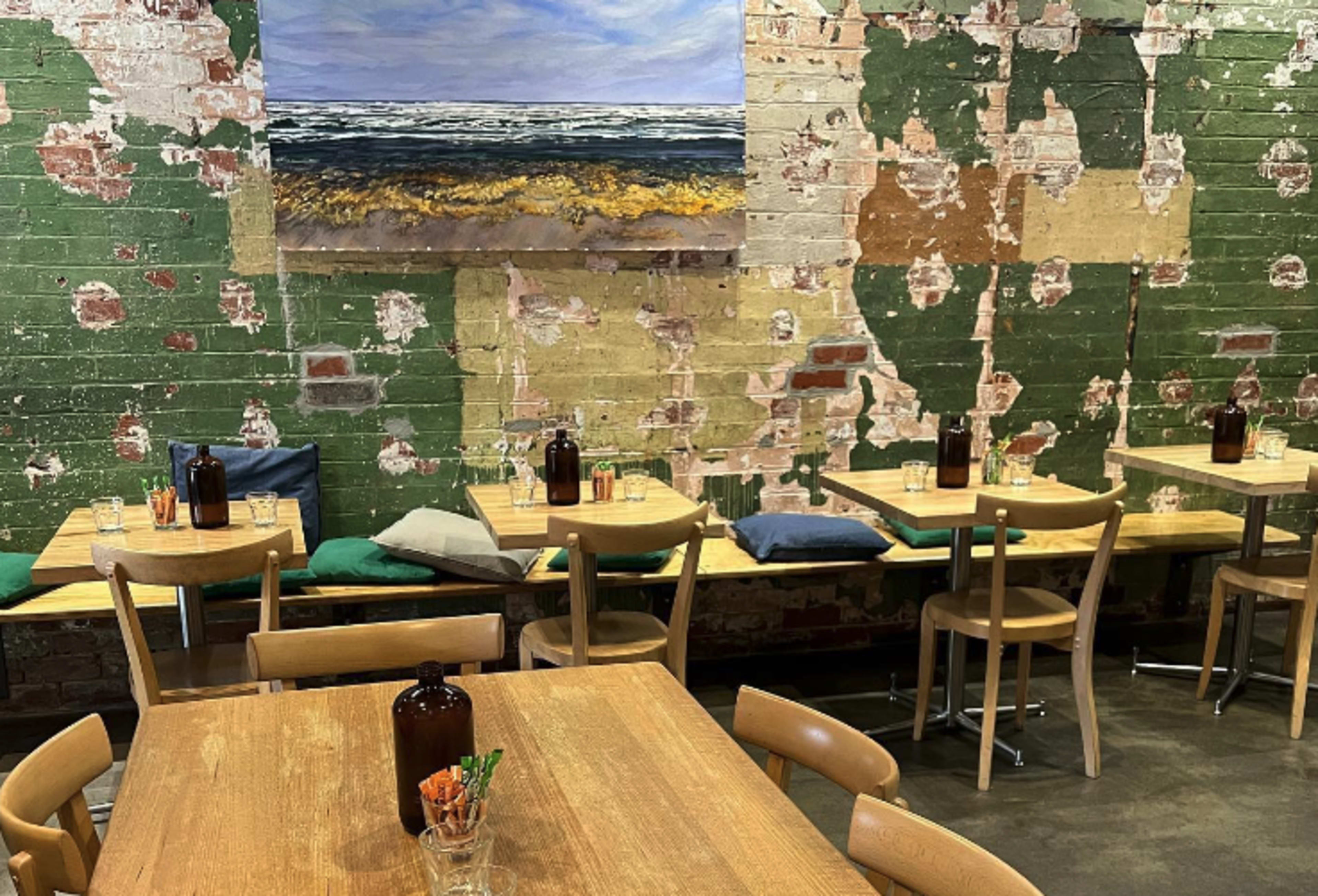 A restaurant interior with wooden tables and chairs, green and beige peeling walls, and an ocean landscape painting hanging above.