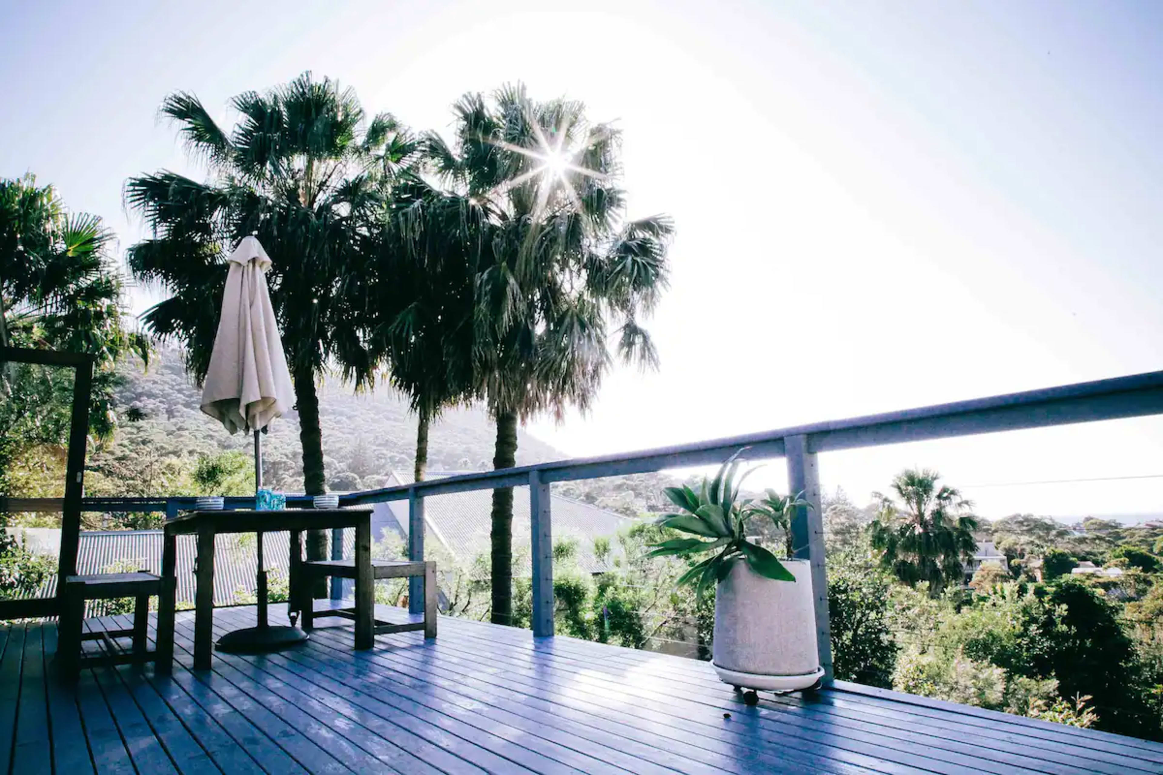 A wooden deck features a table and an umbrella, surrounded by palm trees and a view of nearby hills under a bright sun.