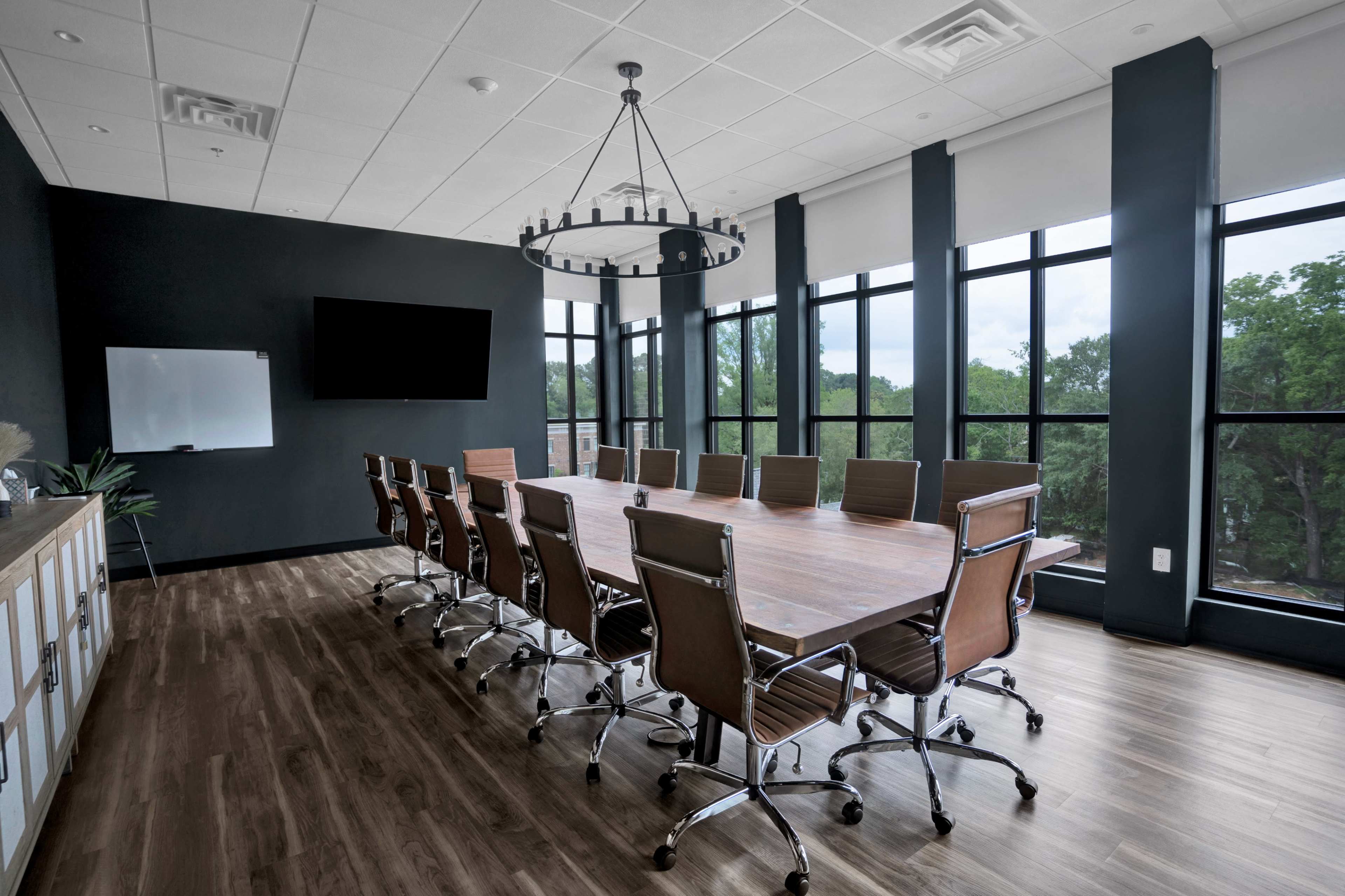A large conference room features a long wooden table surrounded by executive chairs, with floor-to-ceiling windows offering a view of greenery outside.