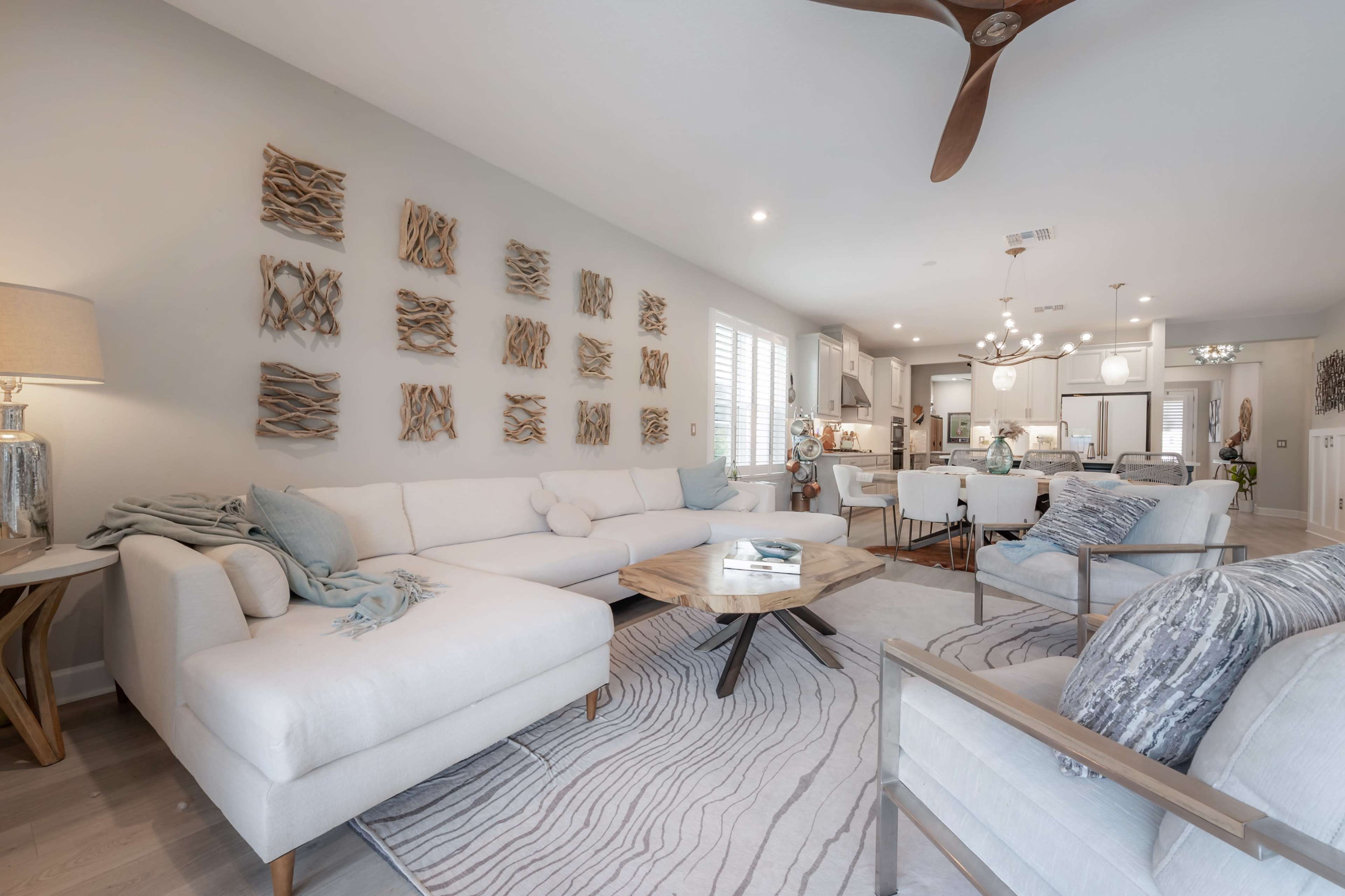 The image displays a modern living room featuring a light-colored sectional sofa, a round wooden coffee table, and decorative wall art, with a view into a kitchen area in the background.