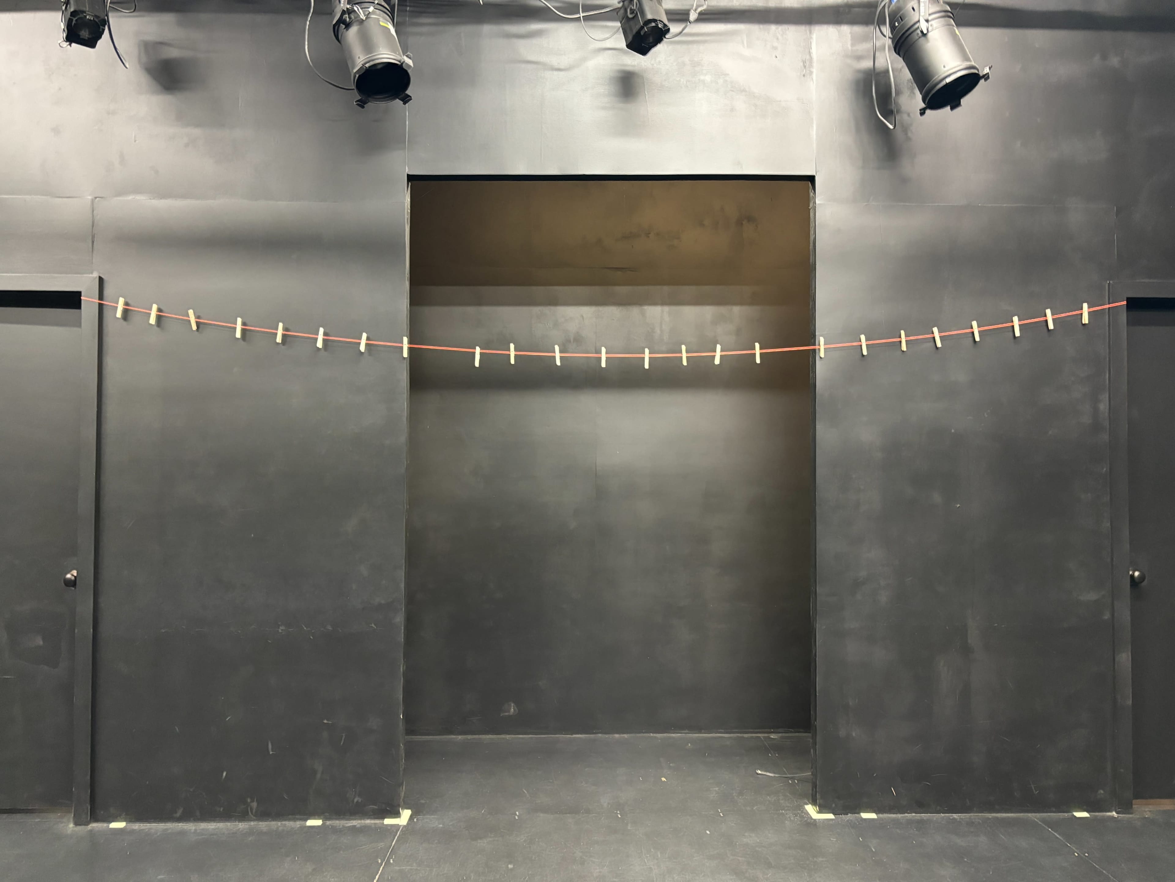 The image shows a stage with a black backdrop and two side doorways, featuring a red line strung across with small clips attached at regular intervals.