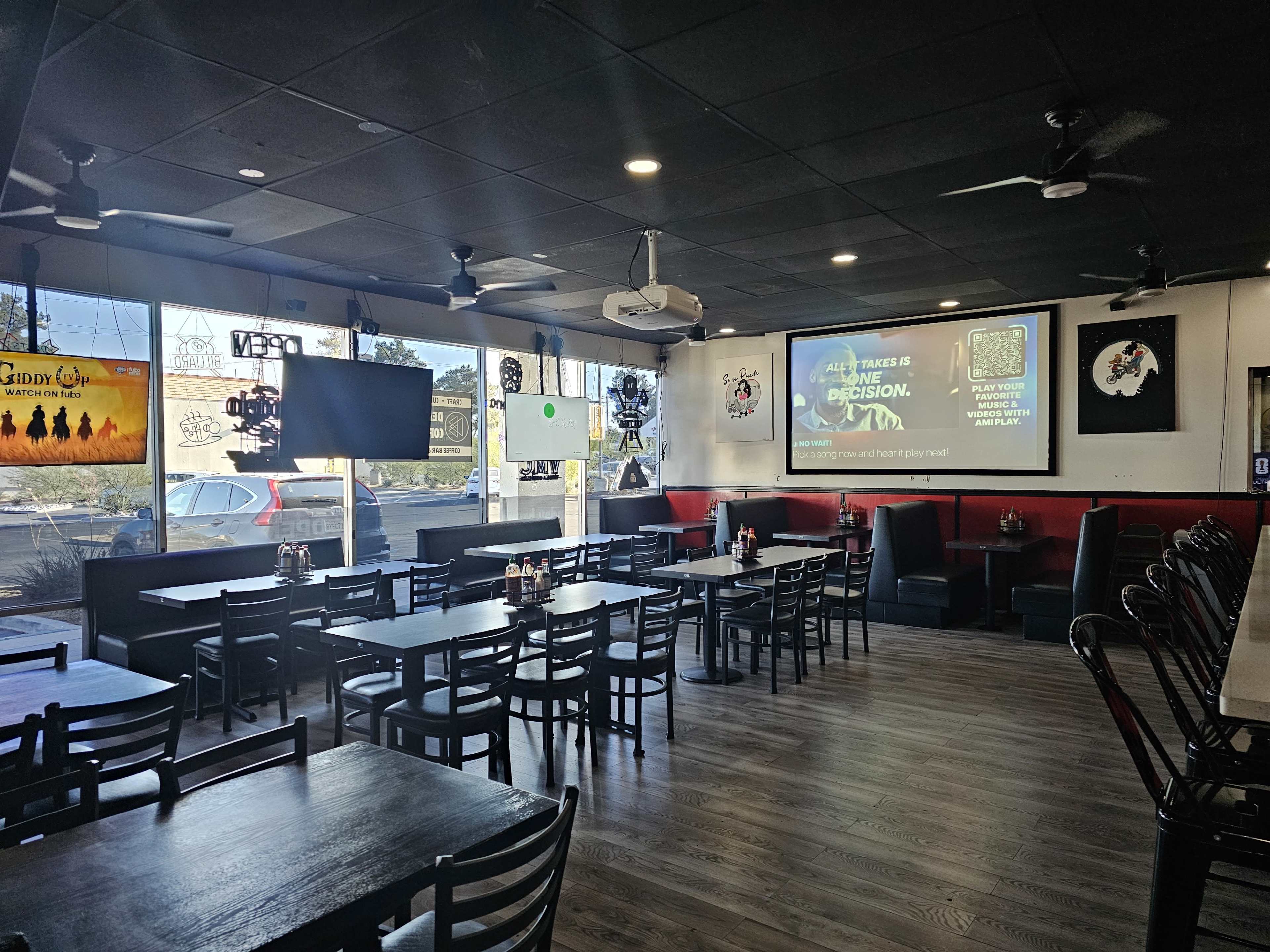 The image shows an empty dining area of a restaurant with rows of black tables and chairs, a large screen displaying a promotional graphic, and a view of the outside through the windows.