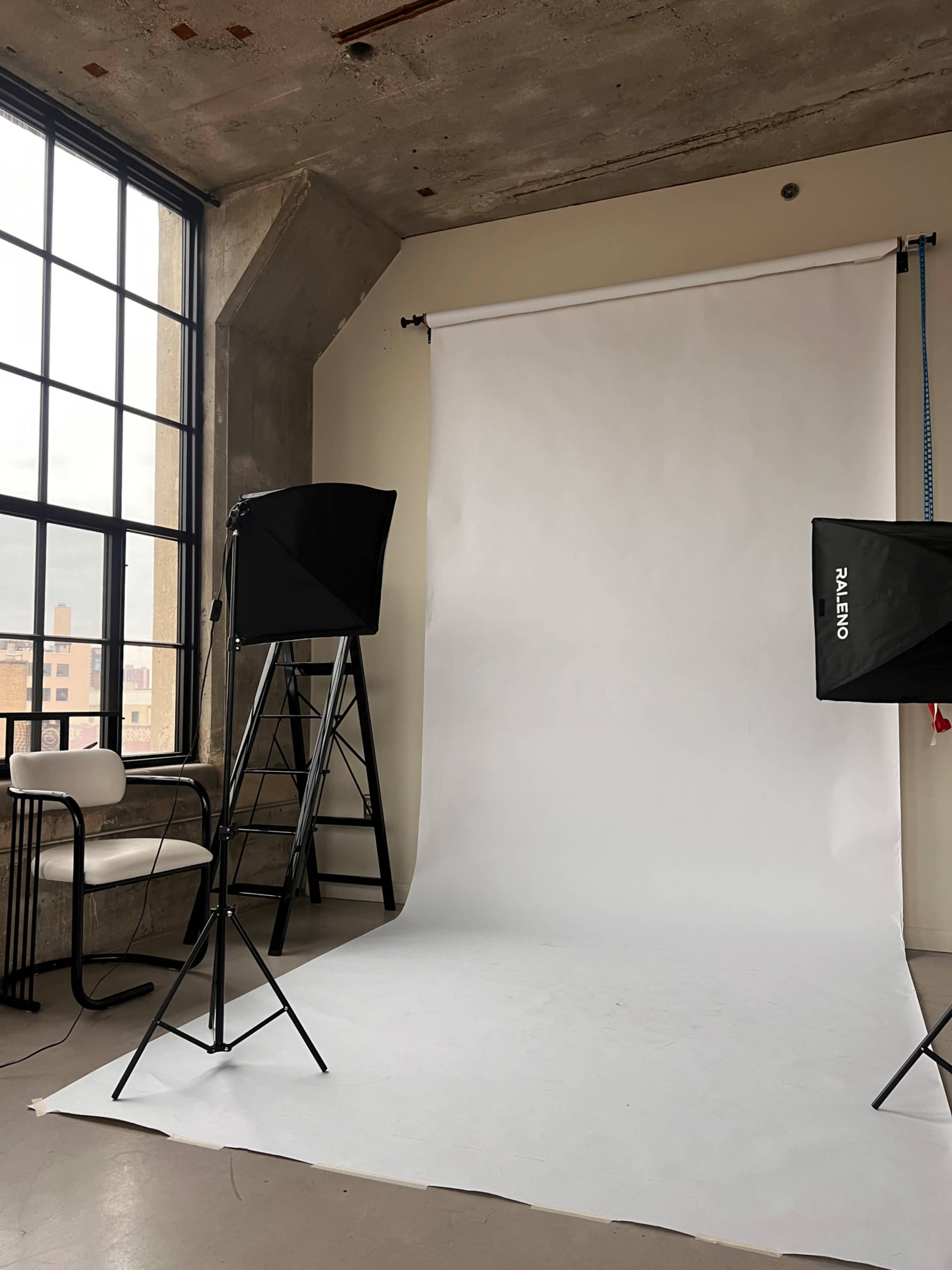 The image shows a photography studio with a white backdrop, two softbox lights, a ladder, and a chair under large windows.