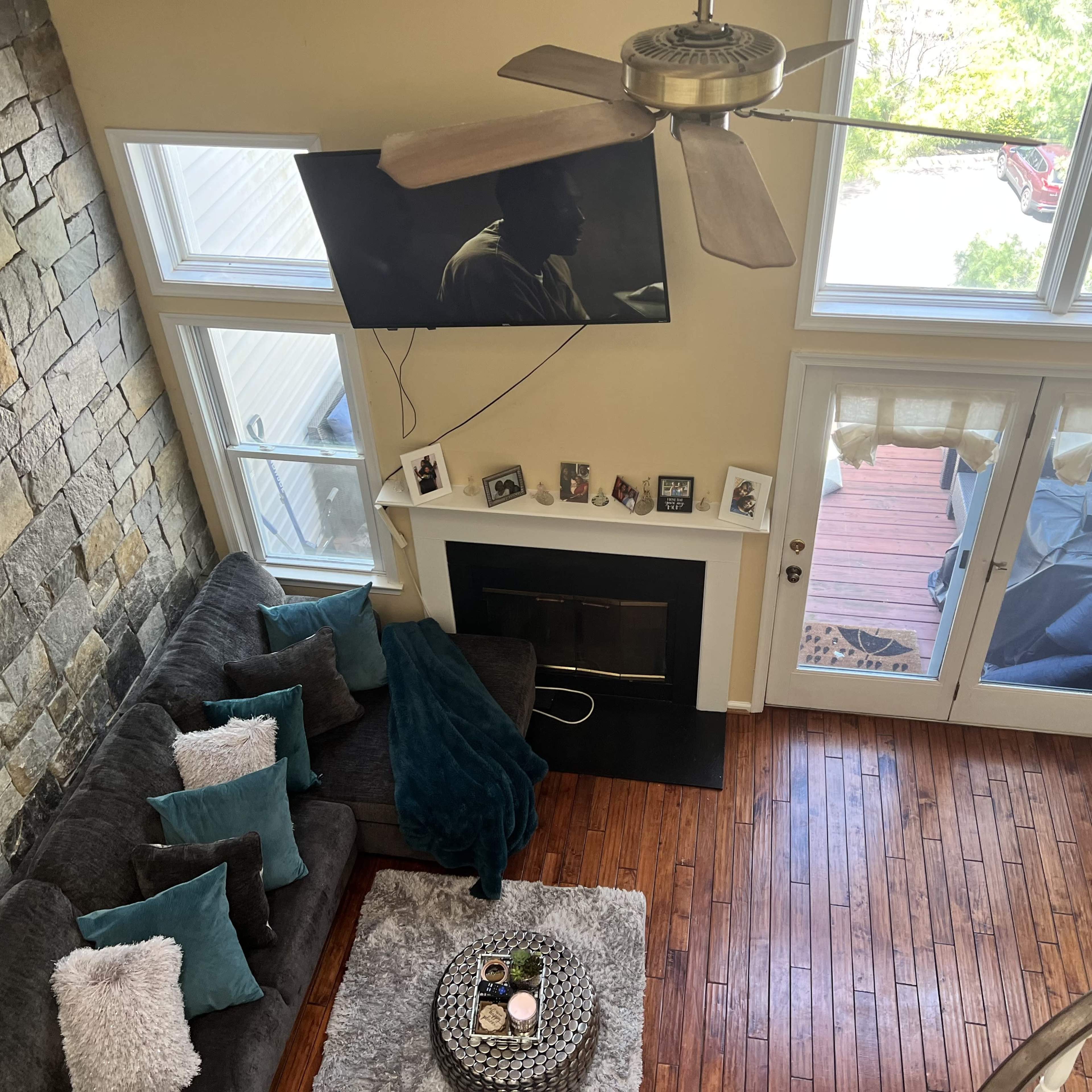 A living room with a stone accent wall, a dark sofa adorned with decorative pillows, a mounted TV, a fireplace, and large windows facing a wooden deck.