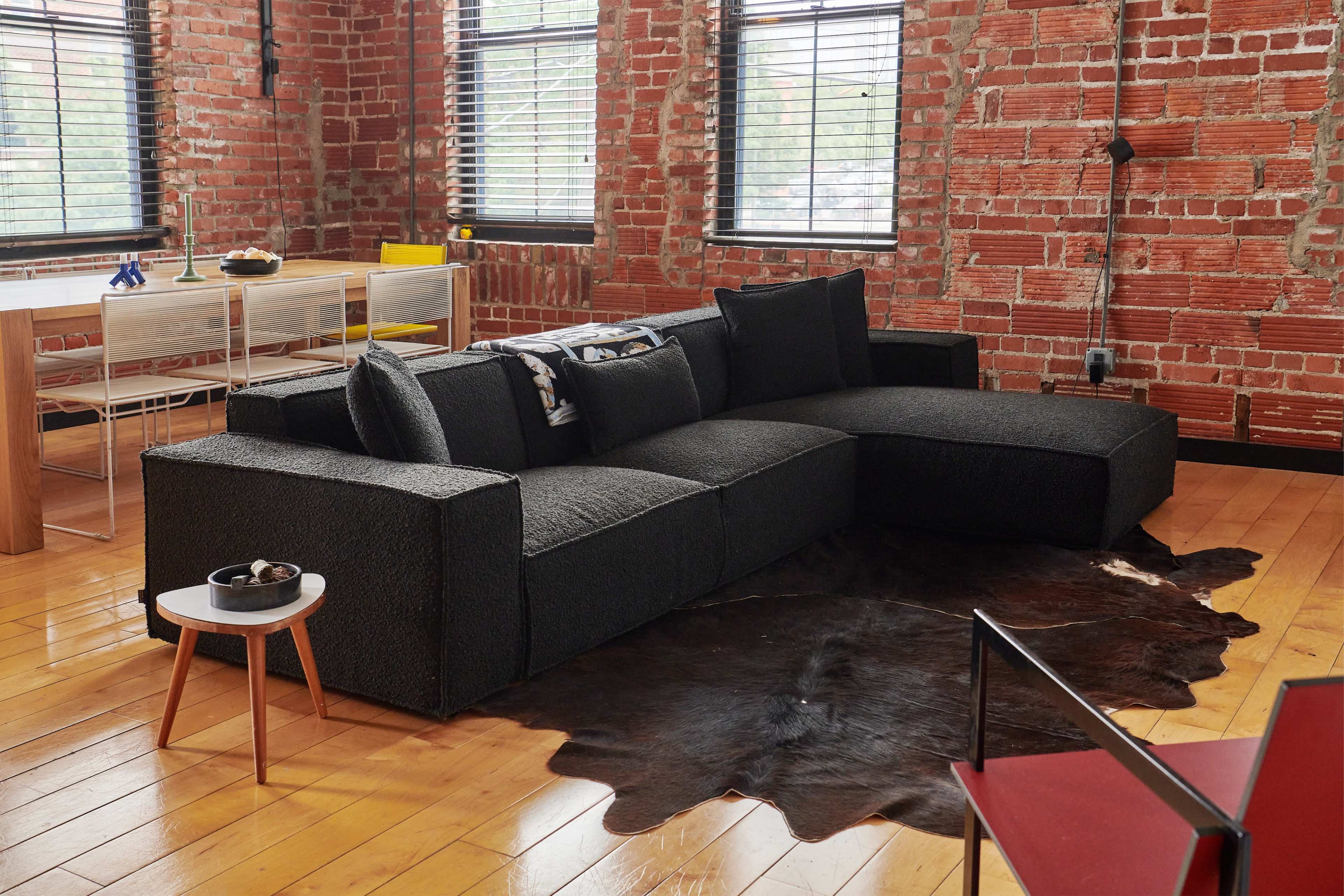 A modern sectional sofa sits on a cowhide rug in a room with exposed brick walls and large windows.