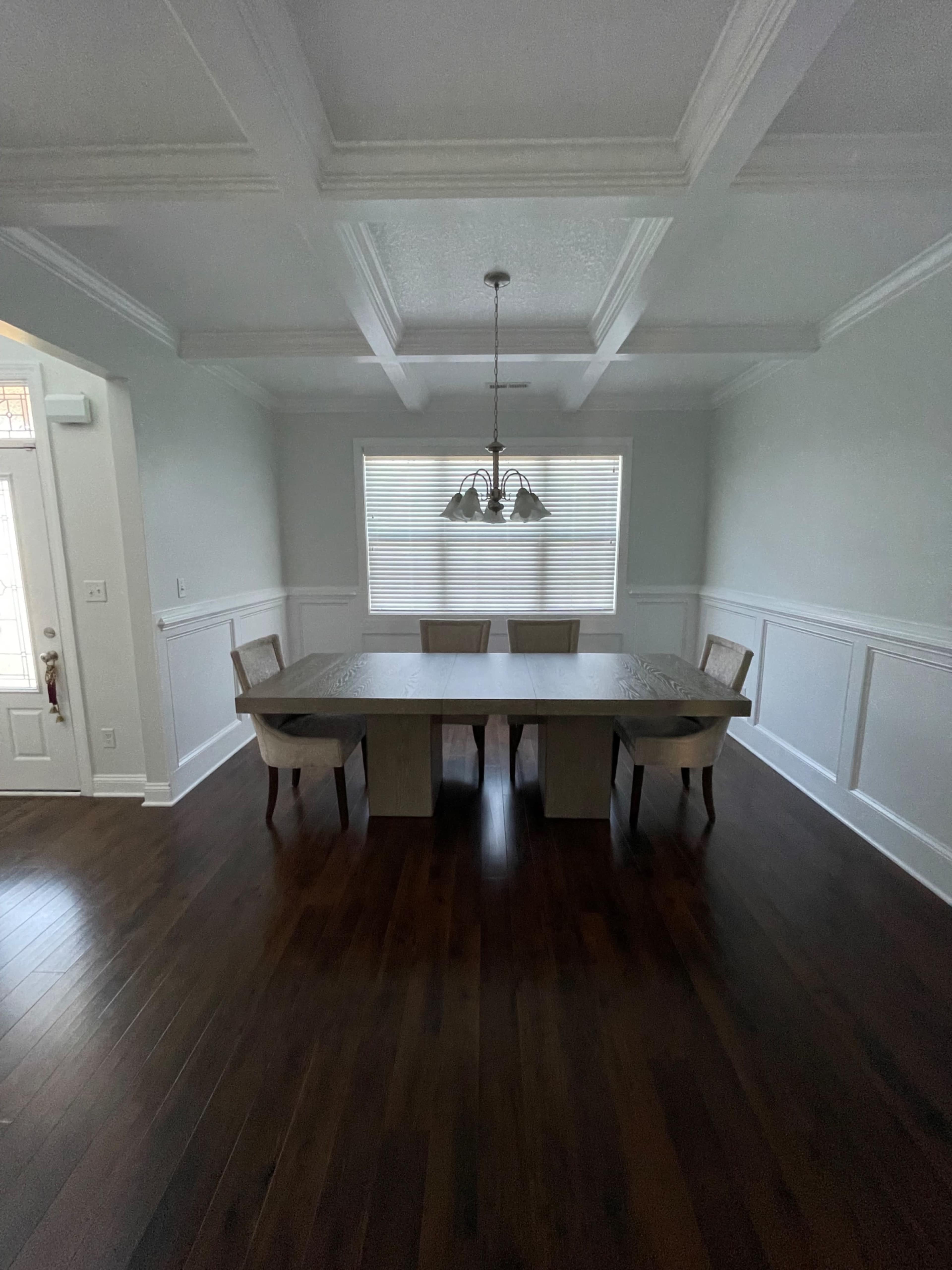A spacious dining room features a long wooden table surrounded by chairs under a multi-light chandelier, with white walls and a coffered ceiling.