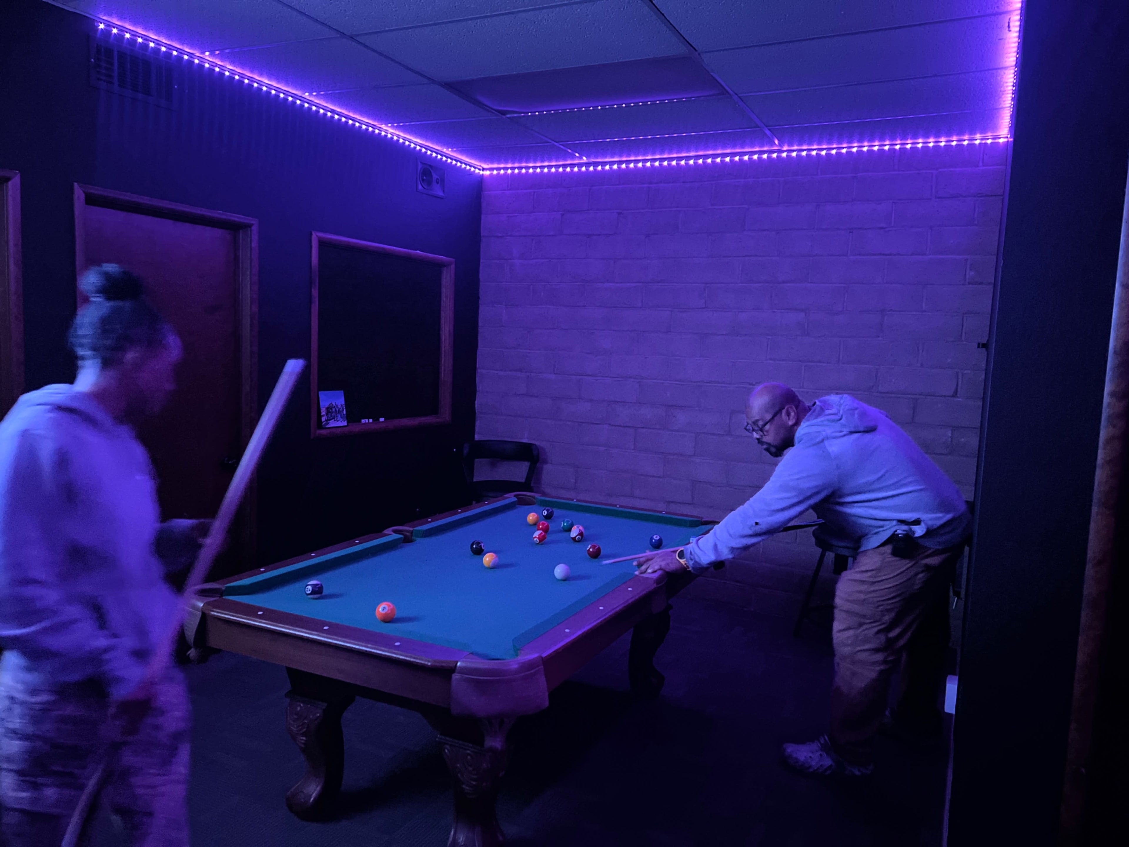 A dimly lit room features a pool table with colorful balls, while two individuals are engaged in a game.
