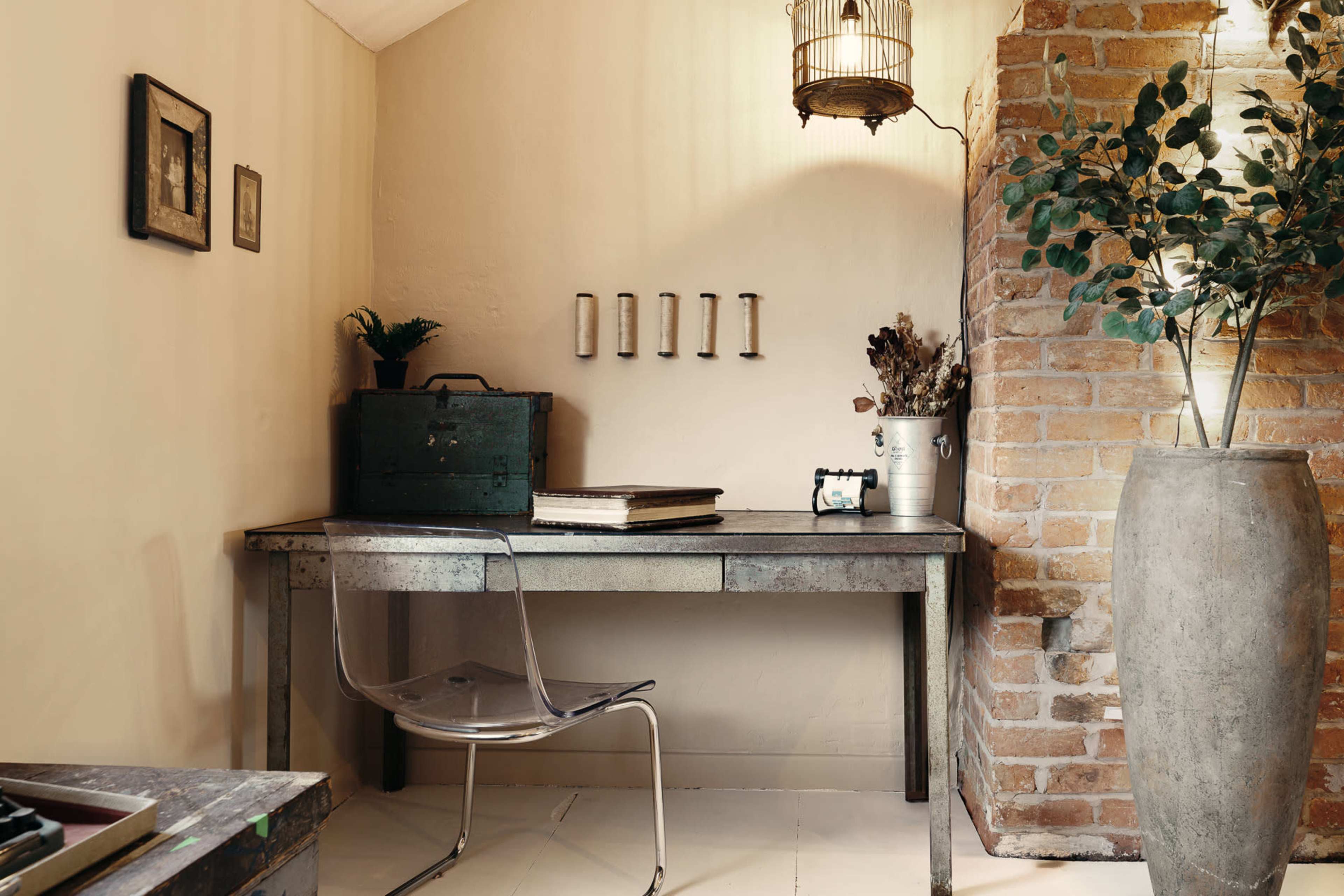 The image features a small, minimalist workspace with a metal table, a transparent chair, and a brick wall adorned with decorative elements and a large vase.