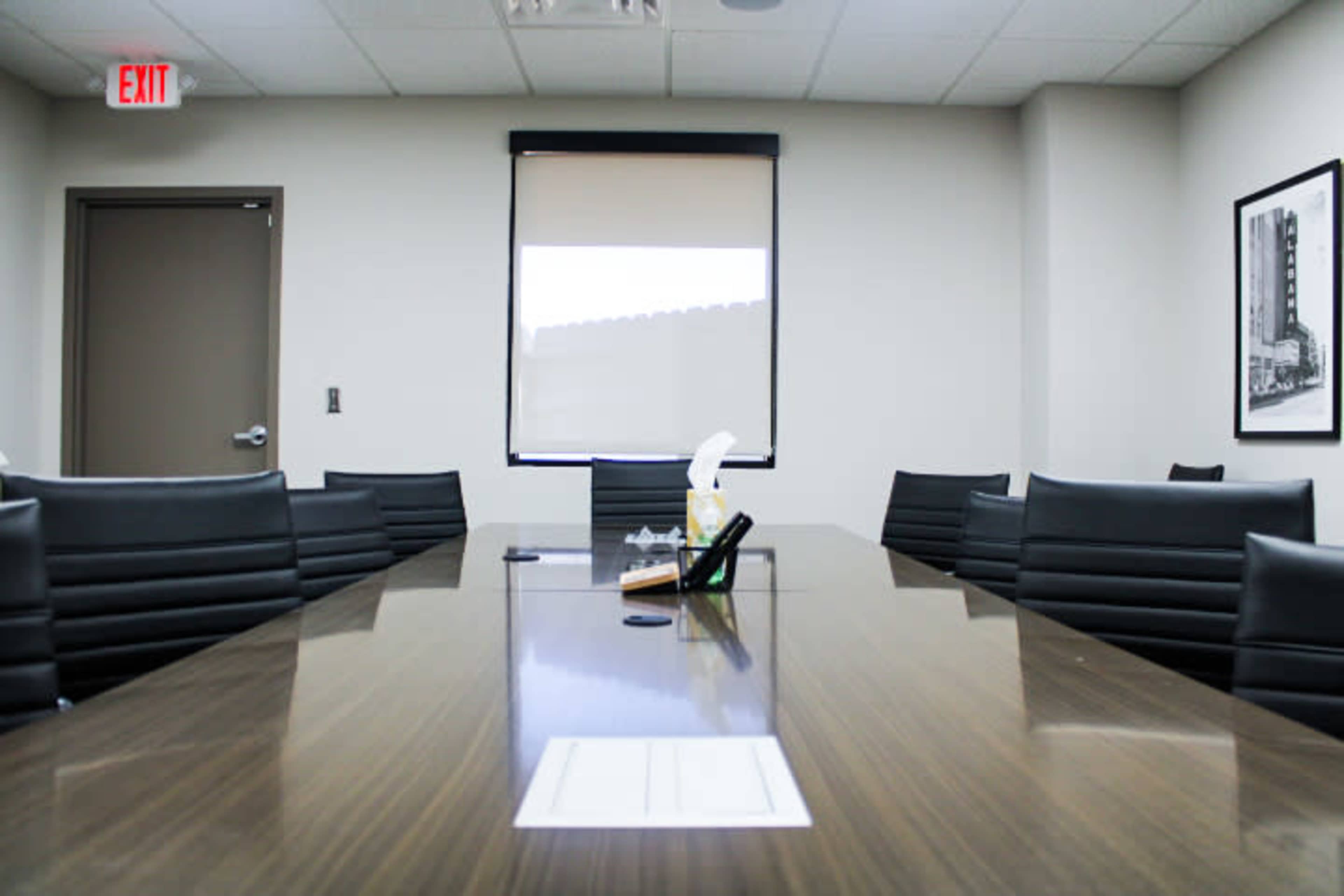 A long conference table surrounded by black chairs is set in a well-lit meeting room with a window and a framed picture on the wall.